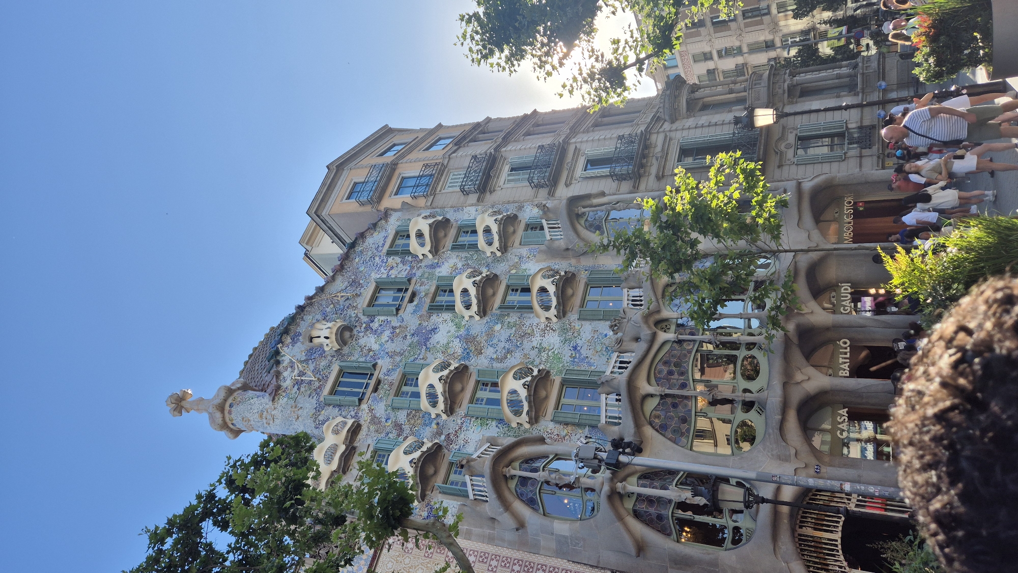 The image captures a sunny day in Barcelona, Spain, with a focus on the Casa Batlló, a renowned architectural masterpiece designed by Antoni Gaudí. The building's facade is a riot of color and undulating shapes, adorned with a mosaic of blue and green tiles and featuring unique, bone-like balconies. Green shutters are visible on many of the windows. To the right of Casa Batlló, a more conventional, multi-story building stands, also with balconies. Trees with lush green foliage frame the scene, partially obscuring the buildings and providing shade.
In the foreground and midground, a crowd of people is visible. Some are walking, while others appear to be queuing or observing the building. A person in the immediate foreground has their head and shoulders in view, looking away from the camera. The street in front of the building has a lamppost and signage indicating "CASA BATLLÓ" and "GAUDÍ," confirming the location. The sky is a clear, bright blue. The overall impression is a vibrant street scene on a pleasant day, showcasing iconic architecture and bustling pedestrian activity.
