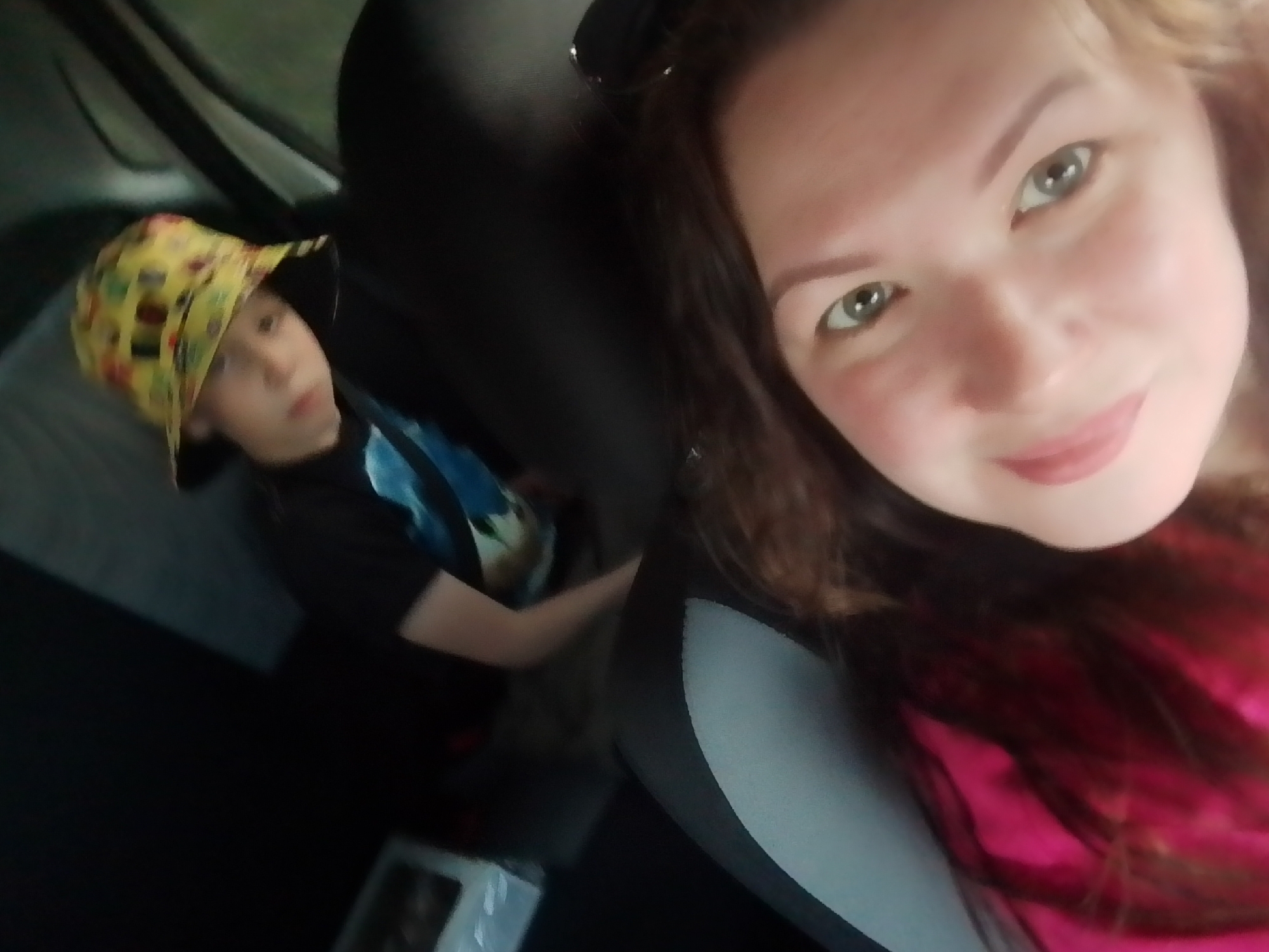 A selfie taken inside a car features a woman in the foreground, occupying the right side of the frame, and a child in the background on the left. The woman has fair skin, green eyes, and light brown hair, and she's wearing a pink top. In the background, the child is wearing a black t-shirt and a yellow hat with colorful patterns. The interior of the car is dark, with the black and gray car seat visible in the foreground. A small, unlabeled box sits in the background near the child. The location is assumed to be the city of Прикамский, Russia.