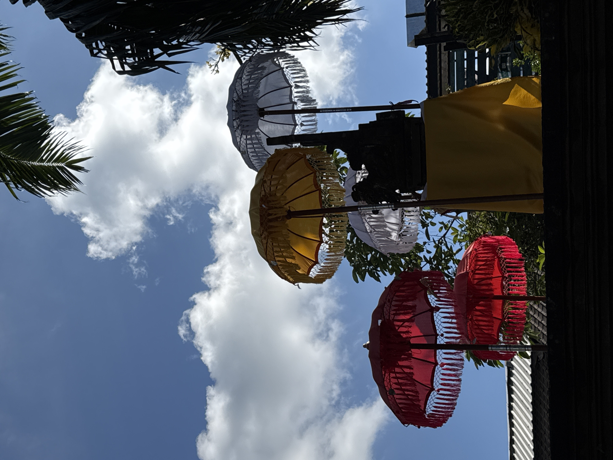 In Ubud District, Indonesia, on a bright, sunny day, several decorative umbrellas are displayed against a vibrant blue sky dotted with fluffy white clouds. The umbrellas, in shades of red, yellow, and white, are ornately decorated with fringe and appear to be part of a cultural or religious celebration. Some of the umbrellas are larger than others and are positioned at different heights. In the background, a silhouetted structure is visible, possibly an altar or a shrine, draped with a yellow cloth. Palm fronds are also visible in the upper right and left corners of the image, suggesting a tropical setting. The scene is captured from a low angle, looking upwards towards the sky and the umbrellas.