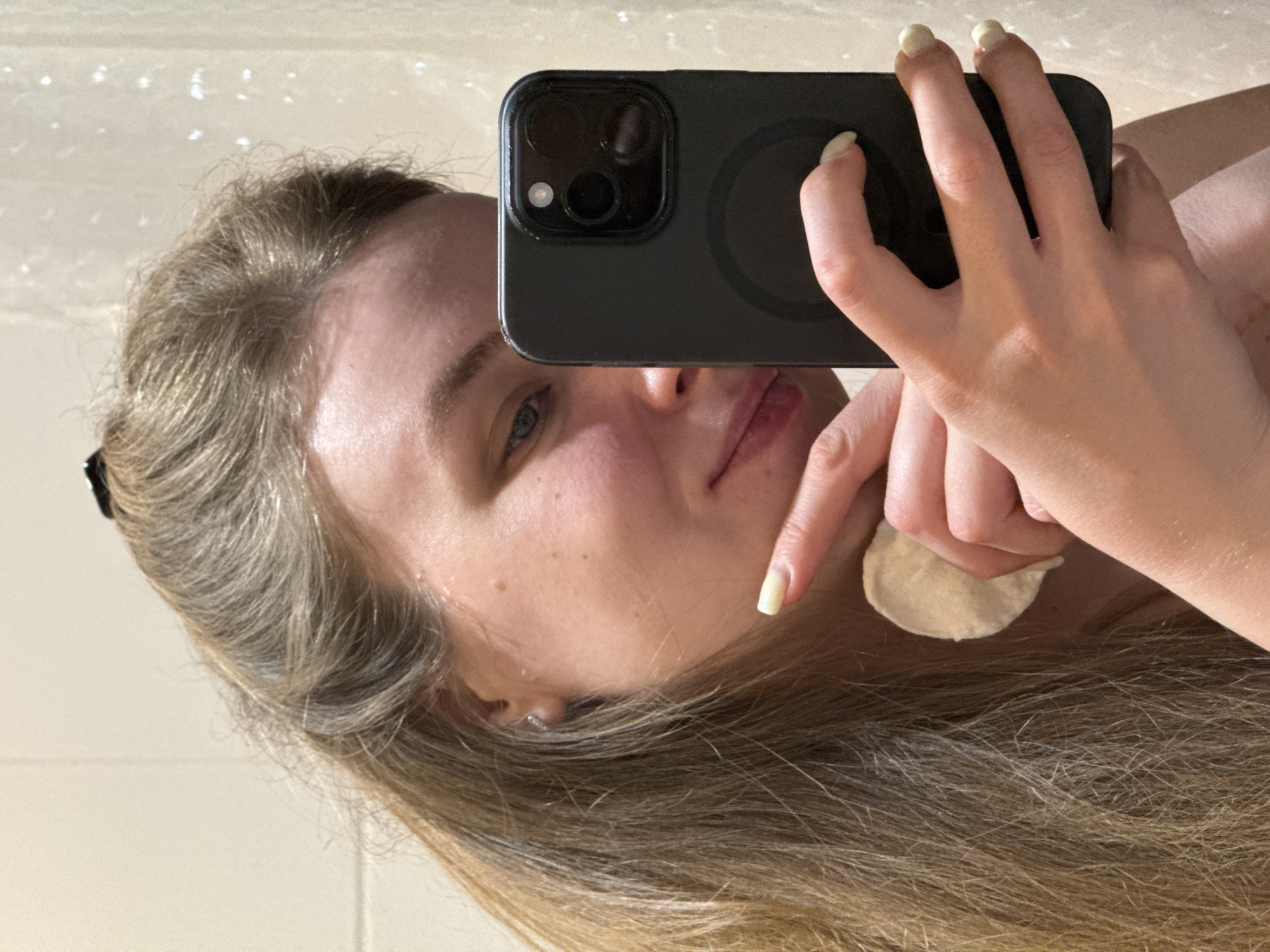 A blonde woman is taking a selfie in a bathroom mirror. She is holding a black smartphone in her right hand, with the camera facing the mirror. In her left hand, she holds a round, white cotton pad near her face. Her hair is long and blonde. She appears to be cleaning or applying something to her face. The background is a plain, light-colored bathroom wall. There is no indication of the location being Vladimir, Russia.
