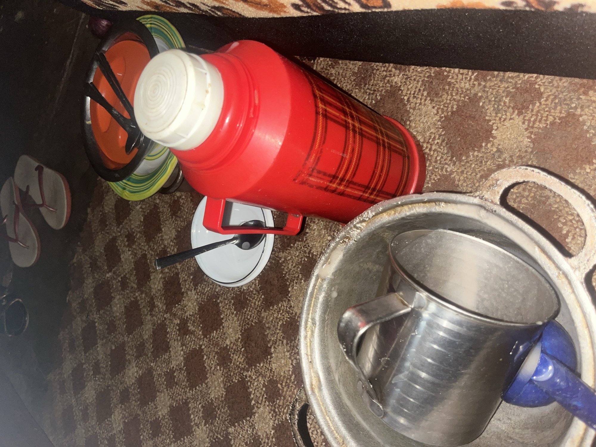 A low-angle, indoor shot depicts a rustic scene on a patterned rug. In the foreground, a large, tarnished metal pot with handles sits open, containing a smaller, shiny metal cup and a blue ladle. To the right, a vibrant red plaid thermos with a white cap stands upright. Behind the thermos, a stack of plates is visible, with two black spoons resting on the top orange plate. In the background, a pair of flip-flops, one red and white and the other white, lies on the floor. A dark, textured surface runs along the right edge of the frame. The overall impression is that of a simple, domestic setting, potentially in Gembu, Nigeria, suggesting a meal or a gathering.