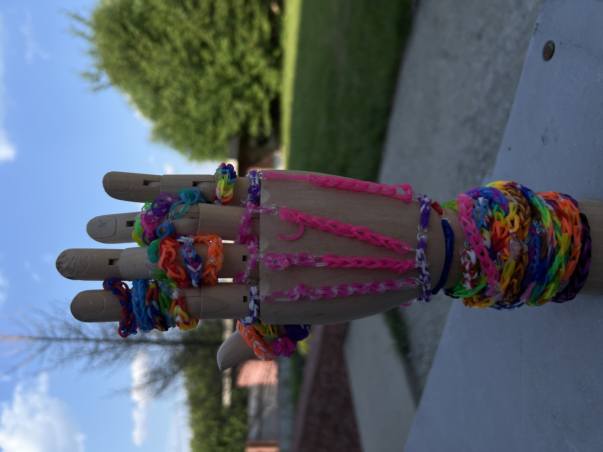 This is a close-up, rotated photograph of a wooden mannequin hand adorned with numerous colorful loom band bracelets. The hand is positioned on a flat, light-colored surface. The bracelets are made from small, elastic rubber bands and are woven into various patterns, creating a vibrant and textured display. Some bracelets are simple bands, while others are more intricate, featuring charms or layered designs. The loom bands come in a wide spectrum of colors, including pink, blue, green, orange, yellow, purple, and rainbow patterns.
The background suggests an outdoor setting. The upper left portion of the image shows a bright blue sky with scattered white clouds, indicating fair weather. A large, out-of-focus green tree is visible behind the hand, and to the right, a blurred green area likely represents more foliage or grass. The lighting suggests it is daytime, likely late morning or early afternoon, with sunlight casting subtle shadows. There is no visible text in the image. The overall impression is one of playful creativity and a celebration of color, likely captured in a backyard or park setting in Dzerzhinsk, Russia, given the provided location context.