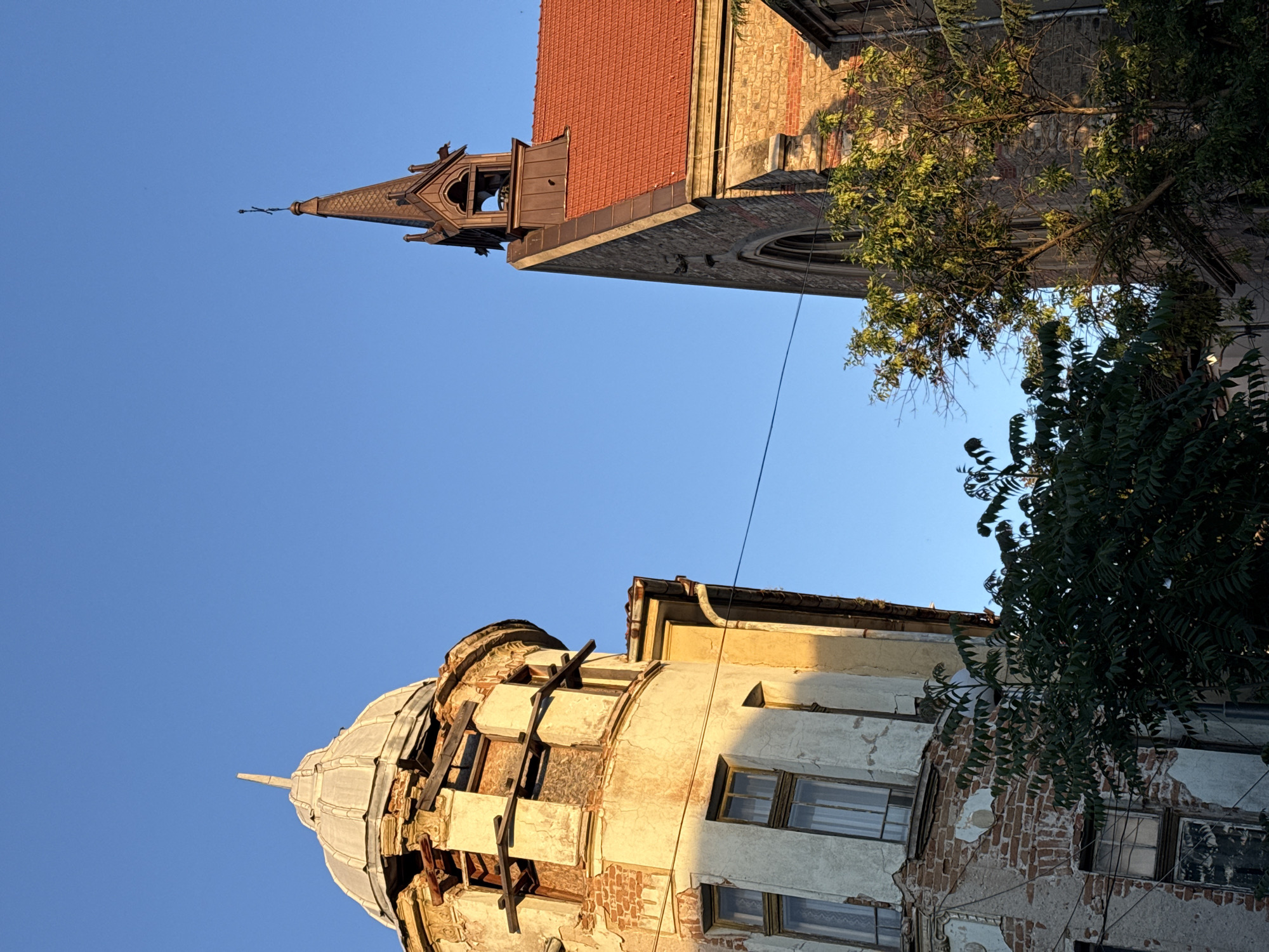 This image shows two buildings against a clear blue sky. The image is taken from a low angle, looking upwards. The building on the top right appears to be a church or cathedral with a red-tiled roof and a pointed steeple. A tree is partially obscuring the building. The building on the bottom left appears to be an older building with a dome-like structure on top. The building shows signs of disrepair, with exposed brick and damaged sections. The buildings are located in the city of Plovdiv, Bulgaria.