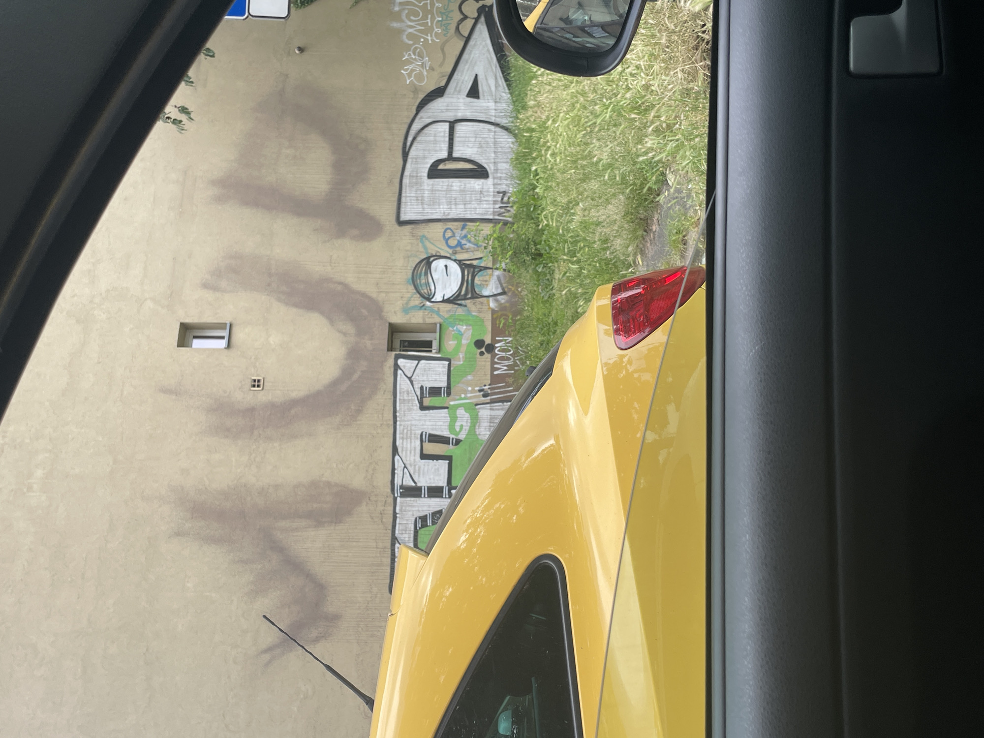 The image, taken from inside a vehicle, looks out at a building facade covered in graffiti. A yellow car is parked on the left side of the frame, obscuring the lower portion of the building. The building's exterior is a light-colored stucco. Large, dark graffiti tags dominate the wall. To the left, faint reddish-brown spray paint forms abstract shapes, possibly remnants of older tagging. To the right, more distinct graffiti is visible, including a stylized black and white "D" and what appears to be a green and black tag with a green outline of what looks like a character. Green foliage grows in front of the building. A small, white-framed window is visible on the building's wall. A side mirror of the car, reflecting the scene, is visible on the right edge of the frame.