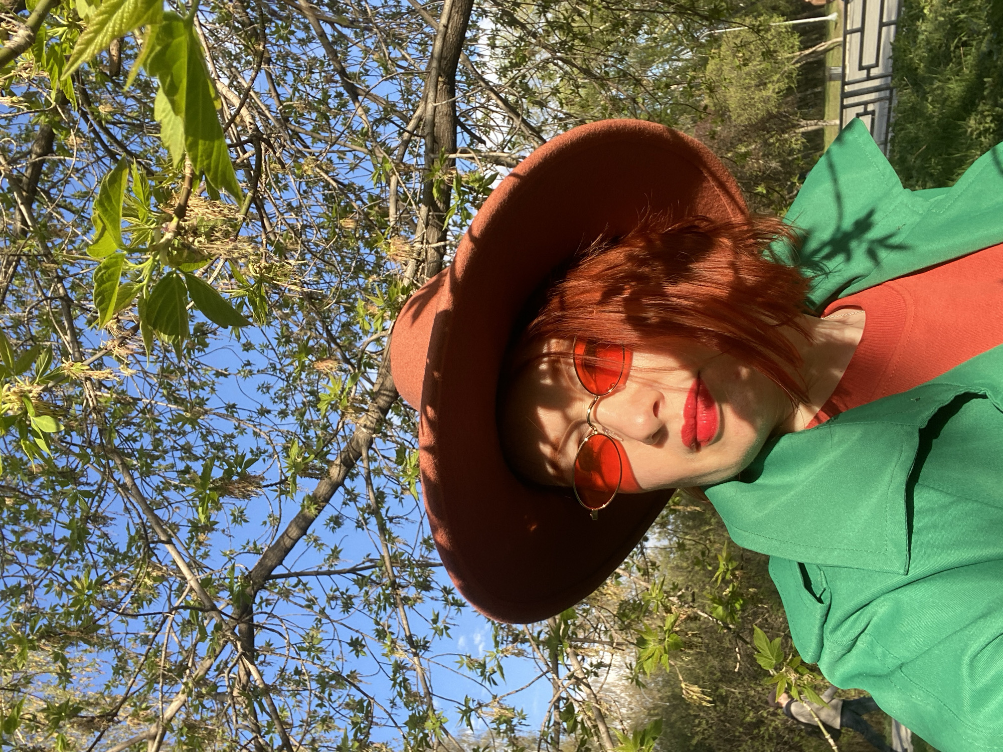 A person is wearing a brown wide-brimmed hat, red sunglasses, a red top, and a green jacket. They are standing outside, surrounded by trees with new leaves in early spring. The sky is visible through the branches. There is a fence in the background. This image was taken in Yekaterinburg, Russia.