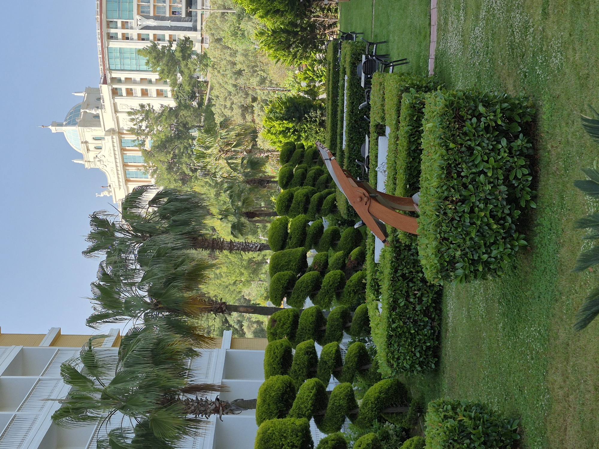 The image captures a scene in Alanya, Turkey. In the background, there is a building with a dome. In the midground, there are several palm trees and sculpted bushes. There is a brown propeller displayed among the bushes. In the foreground, there is a building. The ground is covered in grass. The sky is blue.