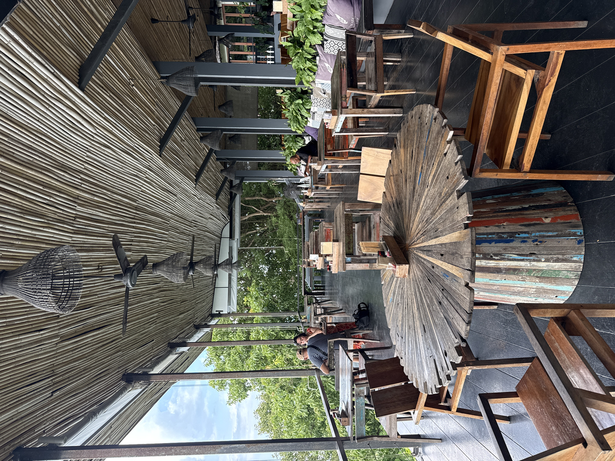 The image depicts an open-air restaurant with a rustic, natural aesthetic, likely located in Ko Samui District, Thailand. The ceiling is constructed from woven bamboo reeds, with several dark, woven pendant lamps and ceiling fans hanging from it. The floor is made of dark wood planks, and metal pillars support the bamboo roof.
Several tables and chairs are arranged throughout the space. In the foreground, a large, circular table with a distressed wooden top, made from numerous small wooden pieces radiating outwards, is prominent. Its base is a cylindrical structure made of vertical, multicolored wooden planks. Wooden chairs with simple, open designs surround this table.
Further back, more tables and chairs are visible, some appearing to be made from reclaimed wood with a weathered finish. One table in the mid-ground has a set of salt and pepper shakers and what appear to be small wooden condiment holders.
There are a few patrons in the restaurant. In the mid-ground, a man wearing a striped shirt and a woman wearing a patterned dress are seated at a table, facing away from the camera and looking out towards the view. Another man, wearing a black shirt, is seated at a table further back on the right, facing a laptop.
The restaurant overlooks a lush green landscape with dense trees, suggesting a natural or tropical setting. The overall atmosphere is relaxed and informal, with a focus on natural materials and a connection to the surrounding environment.
