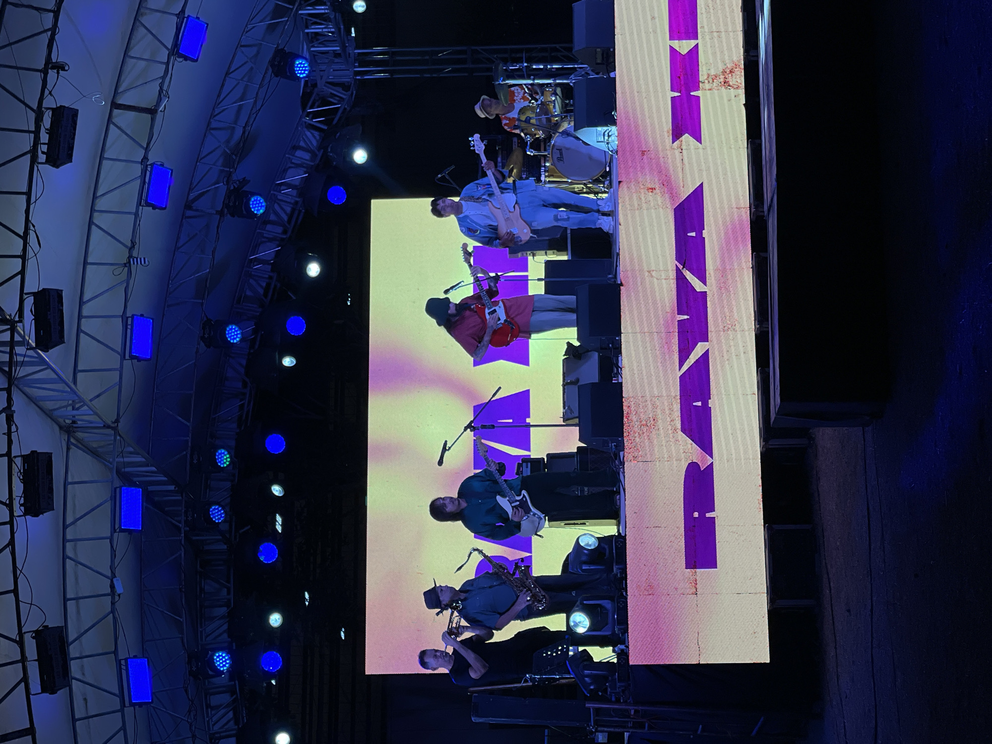 This image shows a live band performing on an outdoor stage at night. The stage is lit with blue and purple lights, and a large LED screen behind the band displays abstract patterns and the text "RAYA KE". There are six band members visible: a trumpeter, a saxophonist, two guitarists, a bassist, and a drummer. The band members are dressed casually. In the foreground, there are speakers and lighting equipment. The background is dark, suggesting an outdoor venue. The event appears to be taking place in Dumaguete, Philippines, as indicated by the text on the screen, which likely refers to a local event or venue. The overall atmosphere is energetic and festive, typical of a live music performance.