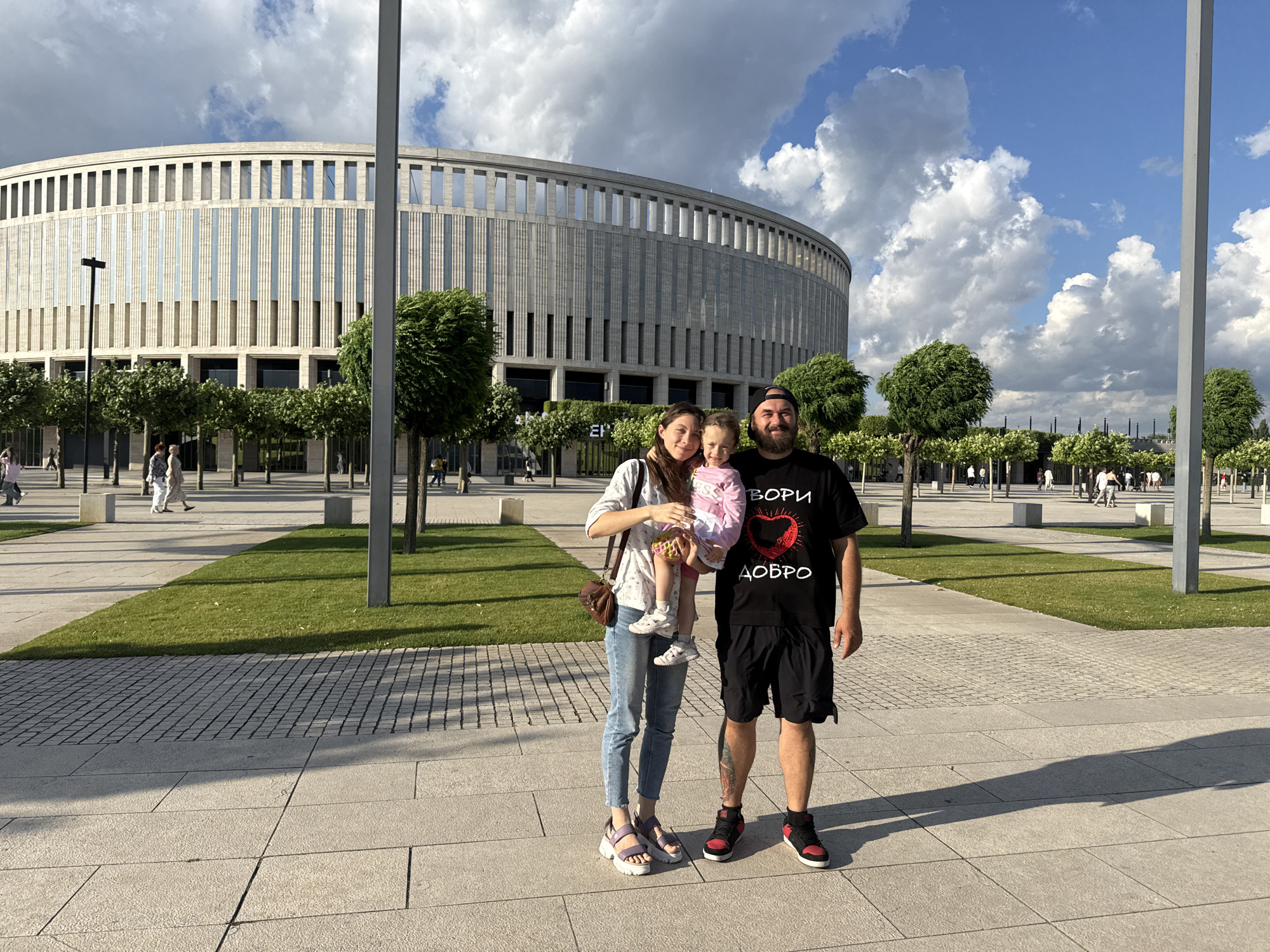 A family of three, a man, a woman, and a young child, are posing for a photo outdoors in front of a large, modern stadium. The man is wearing a black t-shirt with a red heart and text, black shorts, and red and black sneakers. The woman is wearing a light-colored patterned shirt, blue jeans, and light purple sandals. She is holding the child, who is wearing a pink shirt. The stadium in the background is a large, curved structure with many vertical windows and a light-colored facade. In front of the stadium, there are rows of manicured trees and a green lawn. The ground in the foreground is paved with large, light-colored tiles, and there is a grid-like pattern of smaller stones next to the lawn. The sky is blue with scattered white clouds. Several other people are visible in the distance, walking around the area.