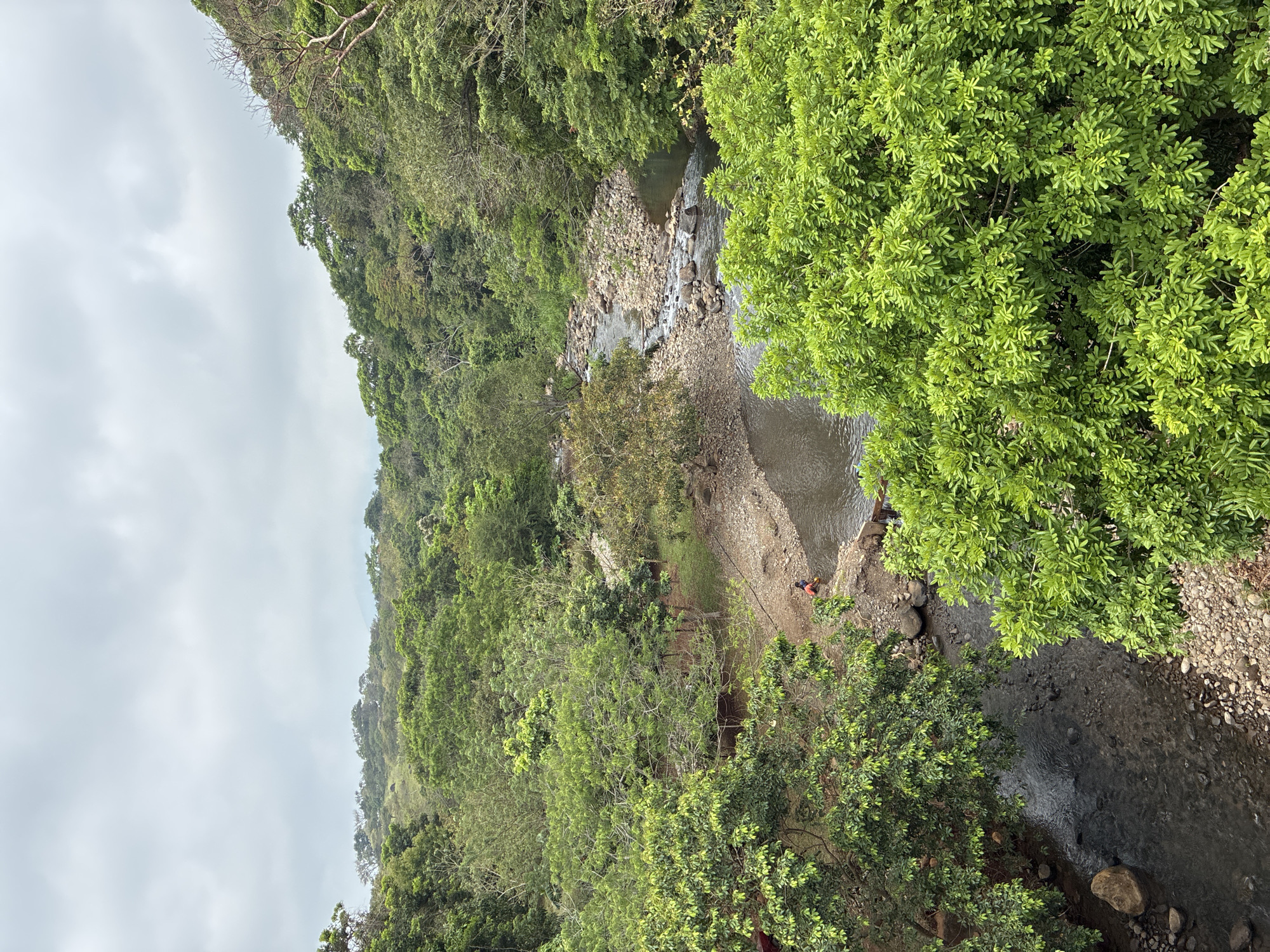 The image shows a view from above of a section of a river or stream winding through a lush, green valley, likely in a rural or natural setting. The location is Pajapan, Mexico. The water appears clear, flowing over rocks and gravel. The banks are lined with dense vegetation, including trees and shrubs. A person or two can be seen on the bank of the river in the middle of the image. The sky is overcast.