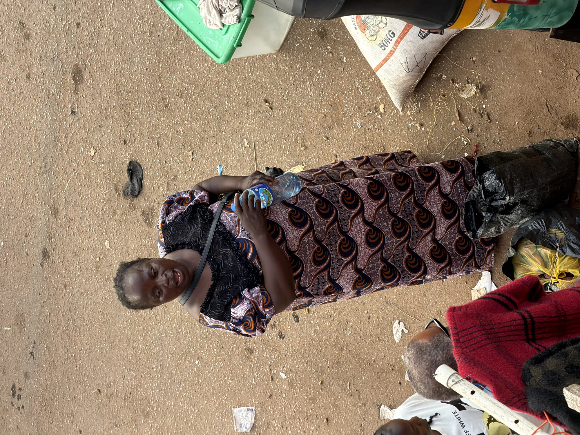 An overhead, medium shot captures a Black woman standing on a dirt ground in Bukuru, Nigeria. She is wearing a patterned dress and holding a bottle of water. The woman has dark skin, short dark hair, and is looking to her left with her mouth open, possibly speaking. She has a black purse slung across her body. The ground is a mix of sand and gravel with some scattered debris, including plastic wrappers and a black sandal. To her right, a green plastic container and a large sack labeled "50KG" are visible, as well as part of a tricycle. In the foreground, to the left, a person wearing a white t-shirt with "OFF WHITE" written on it is partially visible, along with a red and black checkered blanket and a black garbage bag. The scene appears to be an outdoor market or roadside setting.