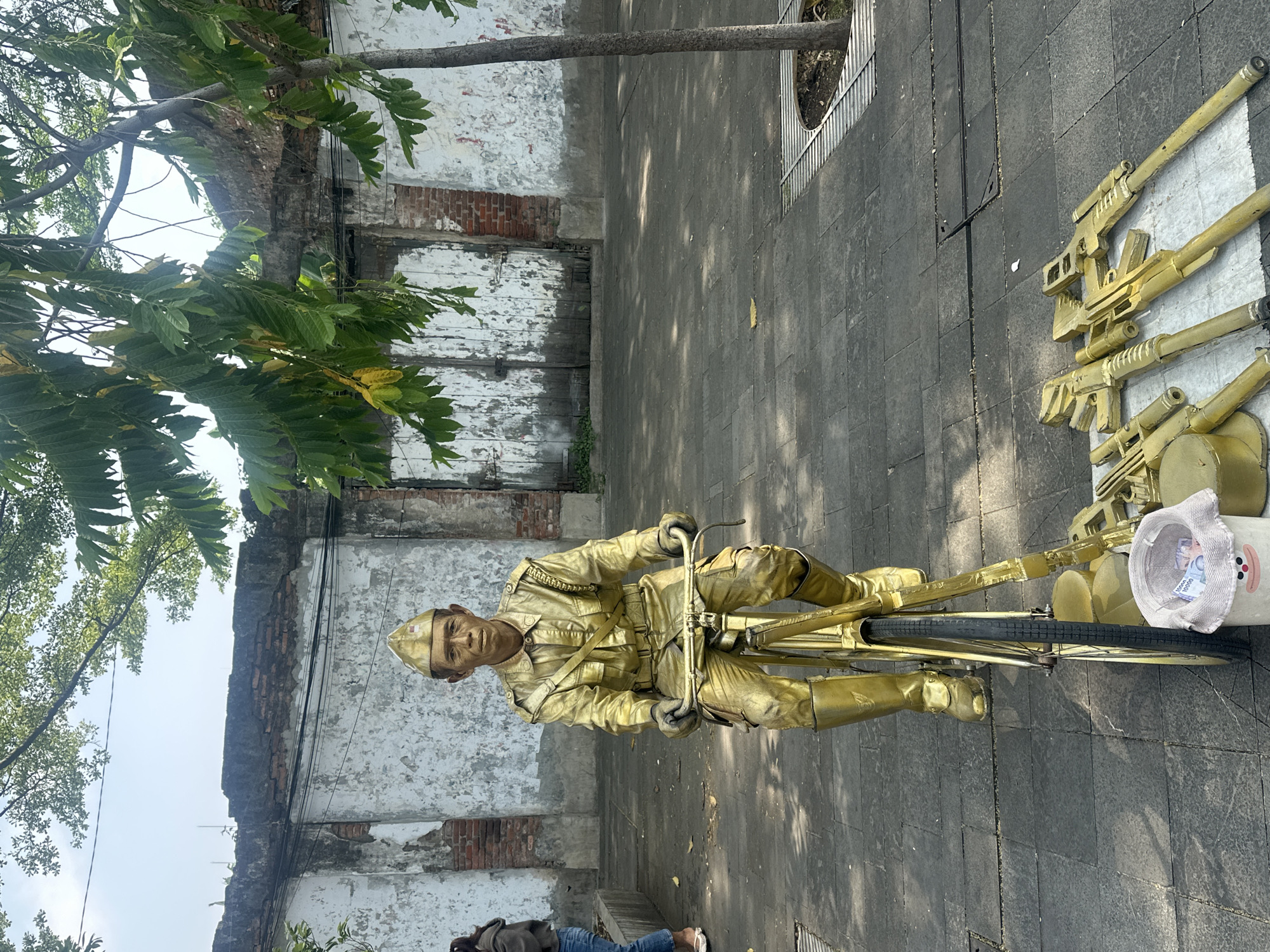 A person painted gold sits on a matching gold bicycle, with golden painted guns on the ground beside the bike. The person is wearing a golden uniform and cap. A white bucket with a smiling cartoon face is positioned near the bike's front tire, containing paper money inside. The scene is set on a gray stone pavement next to a crumbling wall of a building with exposed brick. A tree and foliage overhang part of the wall. This scene is located in West Jakarta, Indonesia.