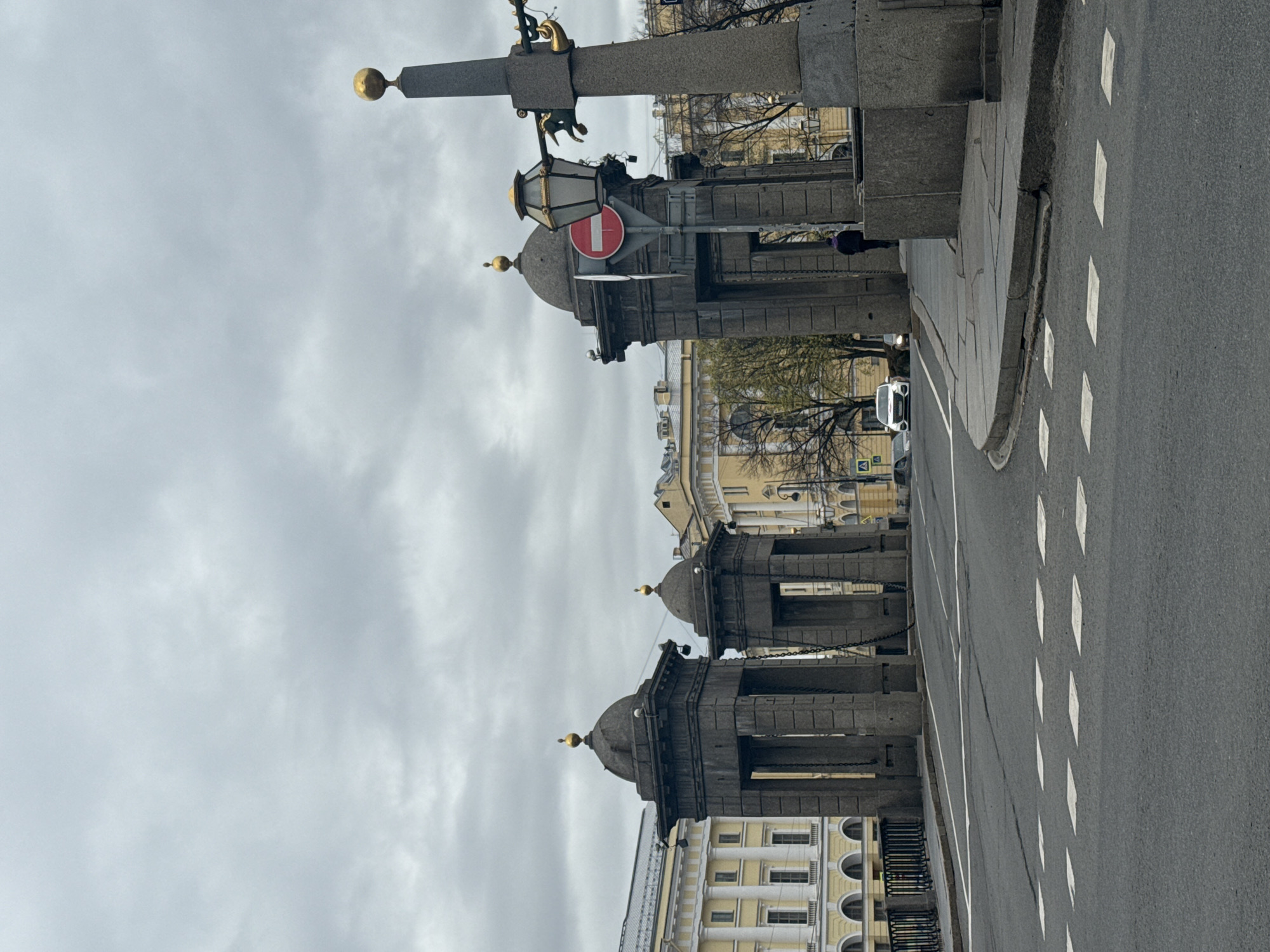 The image shows a street view in Saint Petersburg, Russia, looking down a road lined with gray stone architectural structures. The structures are adorned with gold spheres on top and connected by chains. A "no entry" sign is visible on one of the structures. The road is paved with asphalt and has white lane markings. A white car is visible in the distance. The sky is overcast with gray clouds. Buildings with classical architecture are visible in the background.