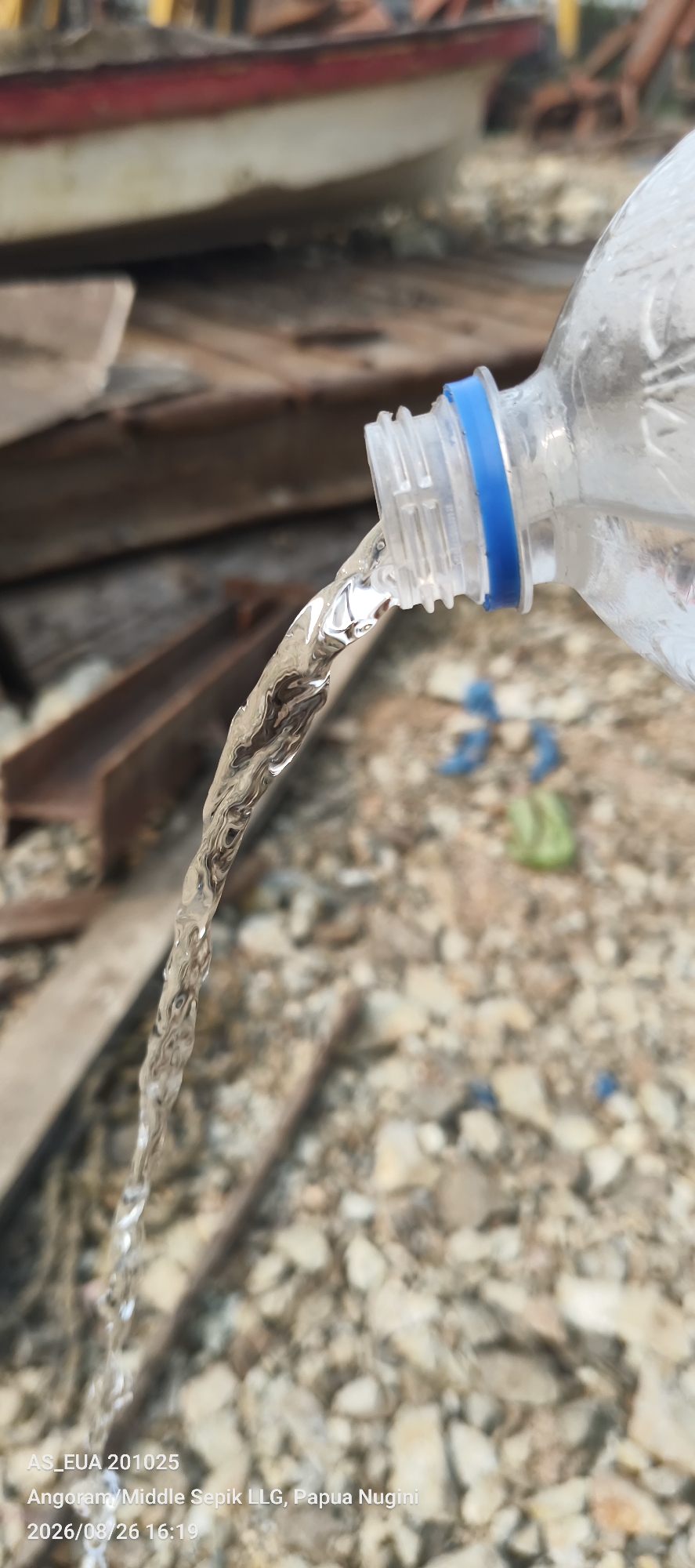 The image shows a close-up of water being poured from a clear plastic bottle with a blue cap onto a surface covered with small rocks and debris. In the background, out-of-focus, are wooden planks and a portion of a red and white structure, possibly a boat or construction material. The water stream is captured in motion, appearing translucent and reflecting light. The ground surface is a mix of light-colored rocks and soil. The text overlay at the bottom indicates "AS_EUA 201025 Angoram/Middle Sepik LLG, Papua Nugini 2026/08/26 16:19", suggesting a location in Papua New Guinea. There are no people visible in the image. The scene appears to be outdoors in an industrial or construction-like environment.