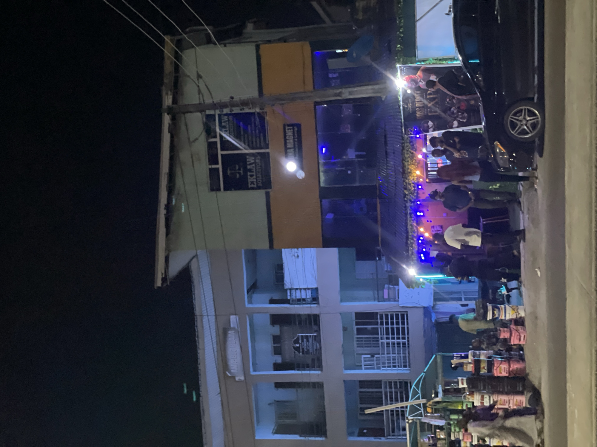 The image depicts a night scene in Ado, Nigeria. A multi-story building is visible with signs for "EKLaw Solicitors" and "Na Magnet Restaurant & Lounge". People are gathered in front of a building, with illuminated blue lights and decorations visible. A black car is parked near the crowd. Stacks of containers, possibly paint or food, are present on the ground.