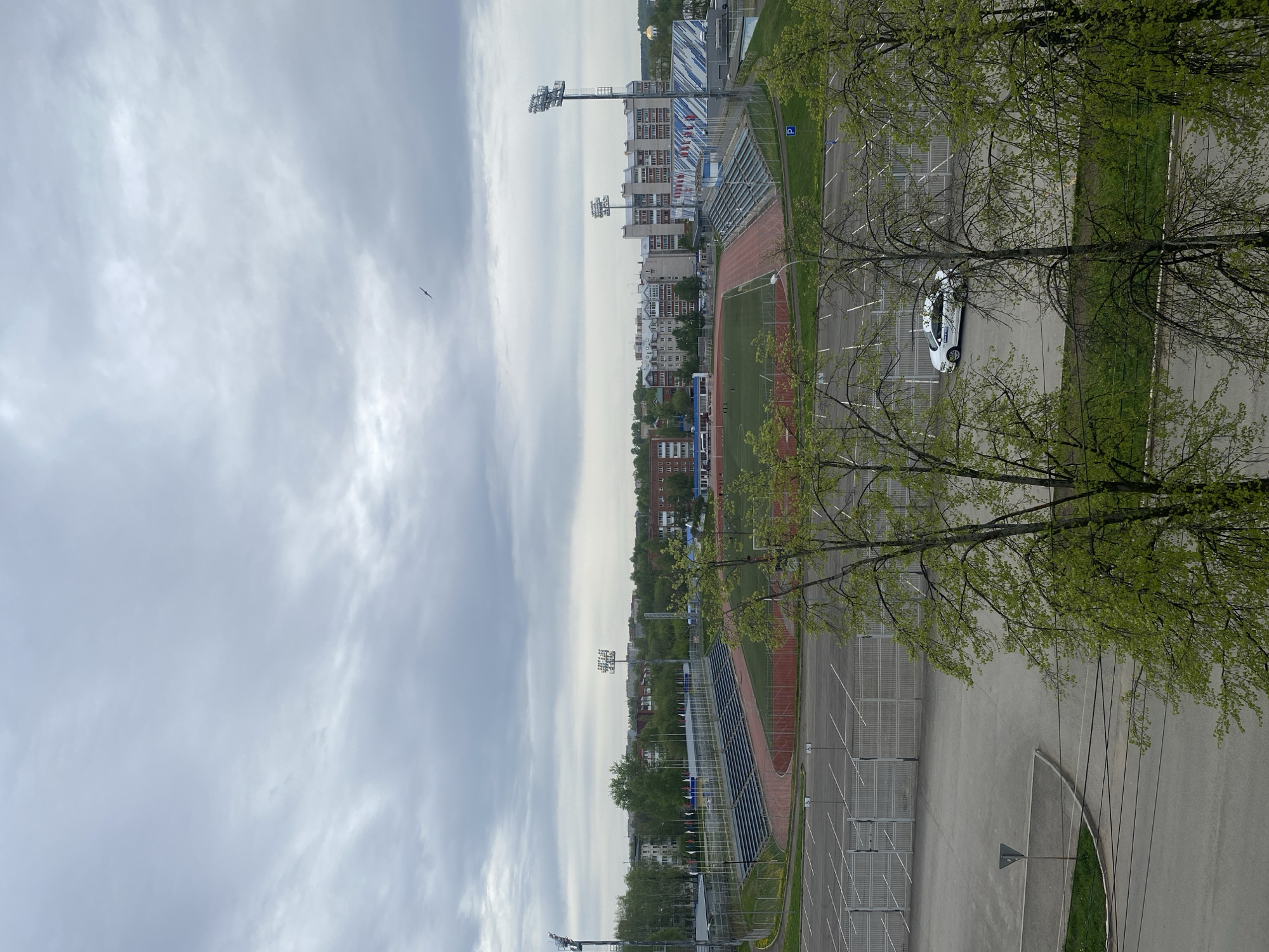 The image shows an overview of a sports complex and surrounding urban environment in Votkinsk, Russia. The sports complex features a stadium with a running track, a green field, and seating stands. Light towers illuminate the field. Adjacent to the stadium is a parking lot, with one white car visible. Trees with fresh spring foliage frame the right side of the image. Beyond the sports complex, the scene transitions to a cityscape with various buildings and structures. The sky is overcast with light gray clouds, and a bird is visible in the sky.