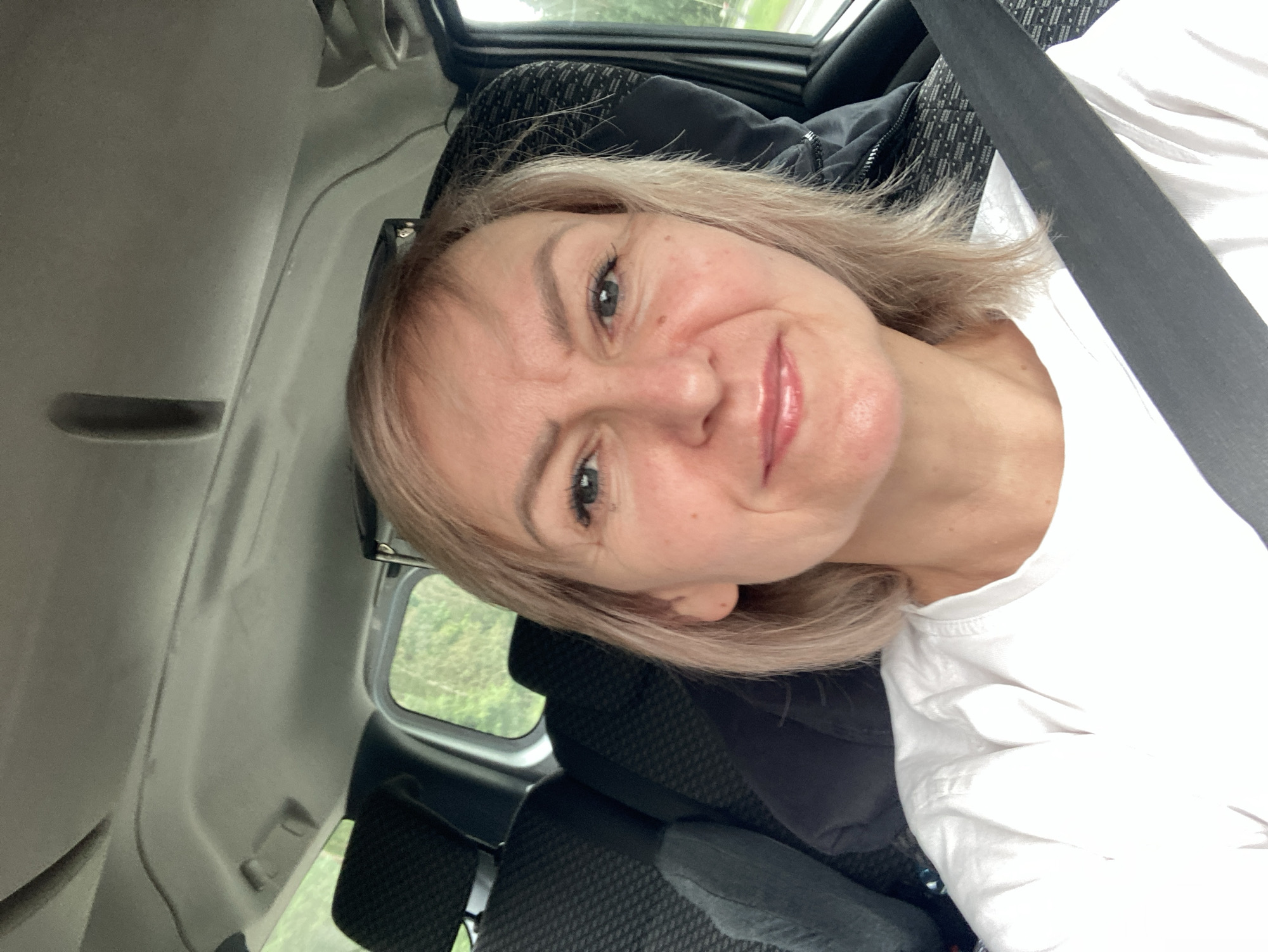 A woman with blonde hair and blue eyes smiles at the camera while sitting in the passenger seat of a vehicle. She is wearing a white t-shirt and a seatbelt is visible across her chest. The interior of the car, including headrests and a portion of the ceiling, is visible. Outside the car window, green trees and foliage can be seen, suggesting a rural or suburban setting. The image appears to have been taken in the vicinity of Драчкаўскі сельскі Савет, Belarus, based on the provided context.