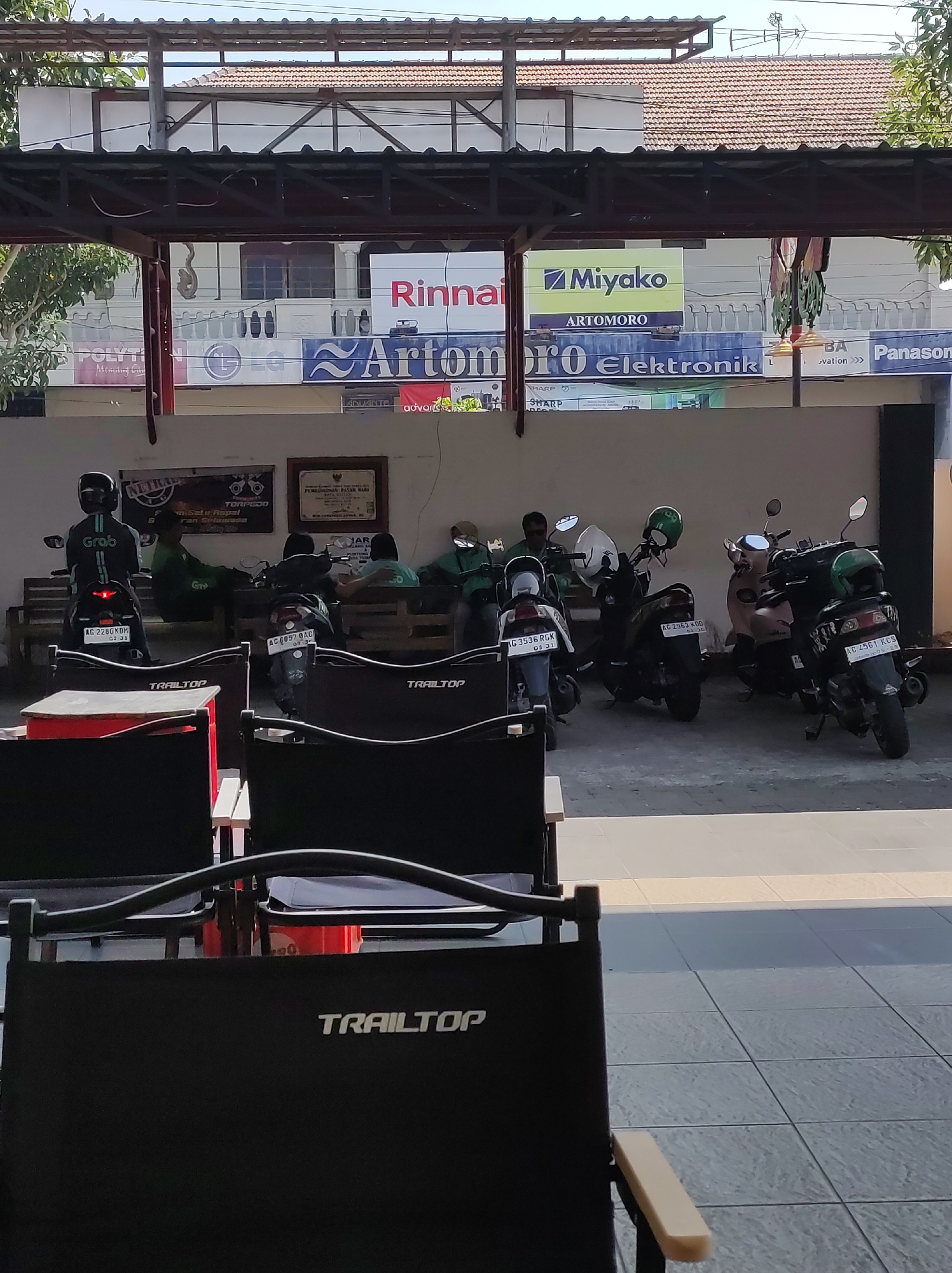 The image shows an outdoor waiting area, likely in Blitar, Indonesia, with several motorcycles parked. In the foreground, two black director-style chairs with "TRAILTOP" branding are visible. Behind the chairs, a row of wooden benches is occupied by several people, most of whom appear to be wearing the green uniform of a ride-sharing service like Grab, indicated by the "GRAB" logo on the back of one person's jacket. Some are sitting and others are standing or interacting. Several motorcycles, including scooters and what appears to be a Grab delivery motorcycle, are parked in a line. The background features a building with signage for various brands, including "Rinnai", "Miyako", "LG", "Artomoro Elektronik", and "Panasonic". The overall scene suggests a public waiting area, possibly at a transportation hub or a commercial establishment. The environment is outdoors with natural lighting, and the architecture suggests a typical Indonesian urban setting.