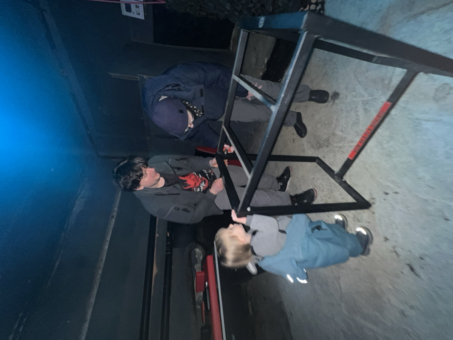 Three people are gathered around a metal structure resembling a jungle gym or a climbing frame. One adult is wearing a blue jacket and beanie, while another wears a dark jacket and a t-shirt. A child in a blue jacket stands near them. The scene is dimly lit, with dark walls or a surrounding structure, possibly indoors. The floor appears to be concrete.