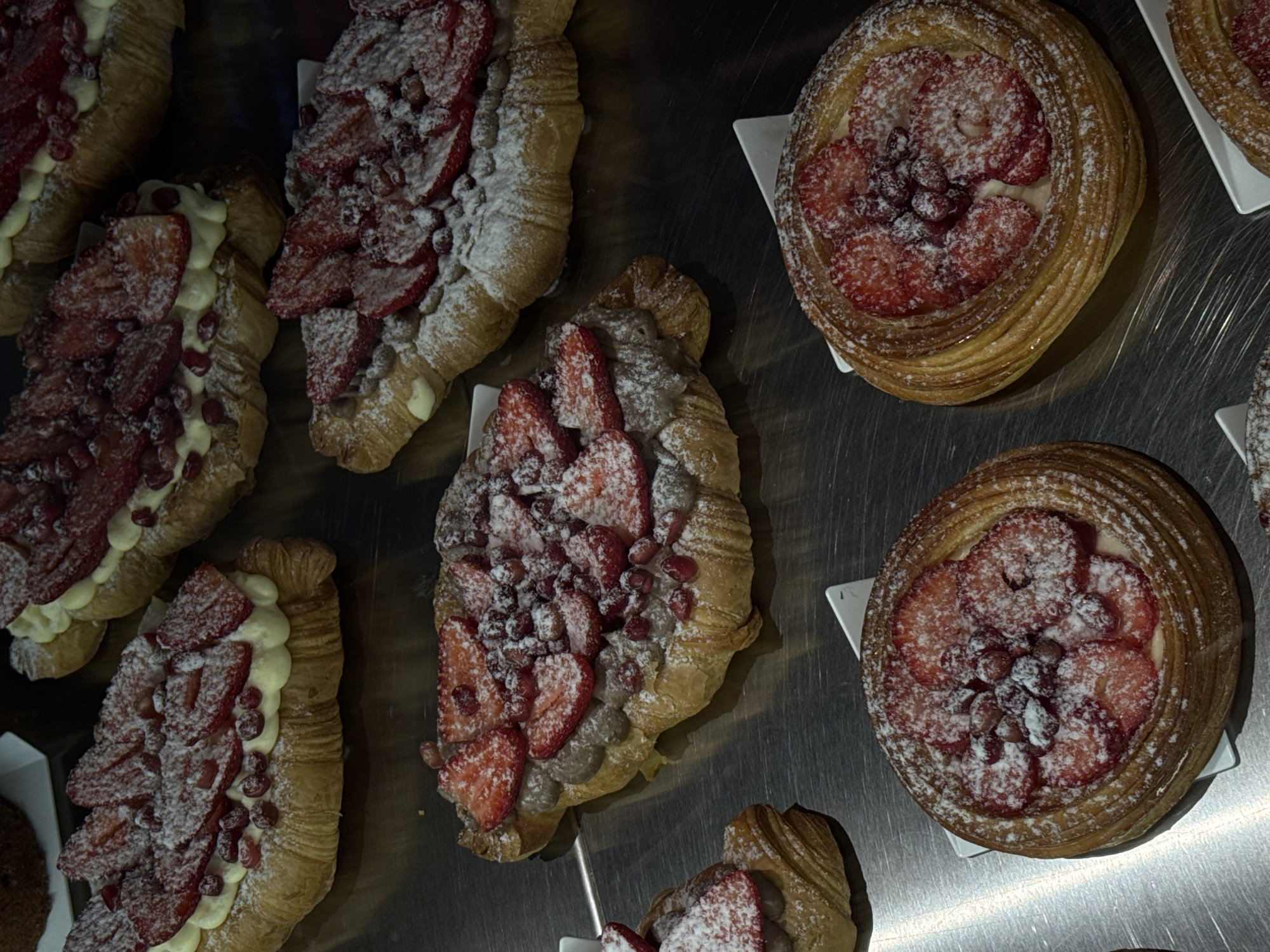 The image presents an inviting overhead view of various pastries, likely displayed on a counter in a bakery or cafe in Yerevan, Armenia. The scene is dominated by an assortment of sweet treats arranged on a shiny, reflective metal surface, possibly stainless steel.
Several elongated, croissant-like pastries are visible, artfully split open and generously filled. These feature a light-colored cream or custard base, topped with slices of fresh red strawberries and scattered dark red berries, which could be cranberries or pomegranate seeds. A liberal dusting of powdered sugar covers the fruit and the flaky, golden-brown pastry, giving them a delicate, frosted appearance. Some of these pastries appear to have a darker filling beneath the fruit, suggesting a variety of flavors.
Interspersed with these are round, layered pastries, reminiscent of Danish pastries or fruit tarts. These individual desserts also showcase a central filling of sliced strawberries and the same dark red berries, all lavishly sprinkled with powdered sugar. Each round pastry rests on a small, white square paper liner, suggesting careful presentation for sale or display.
The lighting is artificial, casting reflections on the metallic surface, enhancing the visual appeal of the desserts. There are no people visible in the frame, and no text is discernible. The overall impression is one of deliciousness and freshness, designed to entice viewers with their vibrant colors and sugary finish.