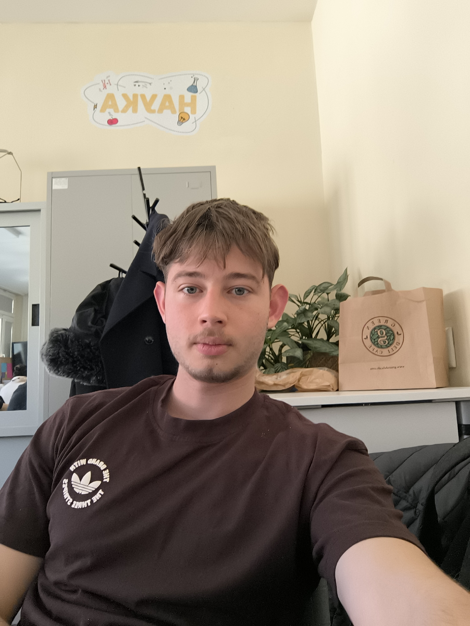 A young man with light brown hair and a neutral expression is featured in a close-up selfie, taken in what appears to be an office or classroom environment in Sofia, Bulgaria. He has short stubble and blue eyes, looking directly at the camera. He wears a dark brown t-shirt with a mirrored Adidas trefoil logo on the chest, surrounded by the reversed text "THE BRAND WITH THE THREE STRIPES".
The background features pale yellow walls and light-colored furniture. To the man's left, a gray cabinet is visible, along with a black coat rack holding a dark coat, potentially with a fur-lined collar. A mirror to the far left reflects a bright room and a window. To his right, a green potted plant sits on a shelf next to a brown paper bag. The bag bears a mirrored circular logo with text, which, when un-mirrored, reads "CREEK DELI," "est. 1130," and "www.thecreekdeli.com."
High on the wall, a cloud-shaped decoration displays a mirrored yellow word "AXYAH," which when un-mirrored and interpreted as Cyrillic, would be "НАУКА" (NAUKA), meaning "SCIENCE." This word is surrounded by small, stylized illustrations related to science and ideas, including a DNA strand, an atom, a test tube, an apple, and a lightbulb. The lighting suggests it is daytime.