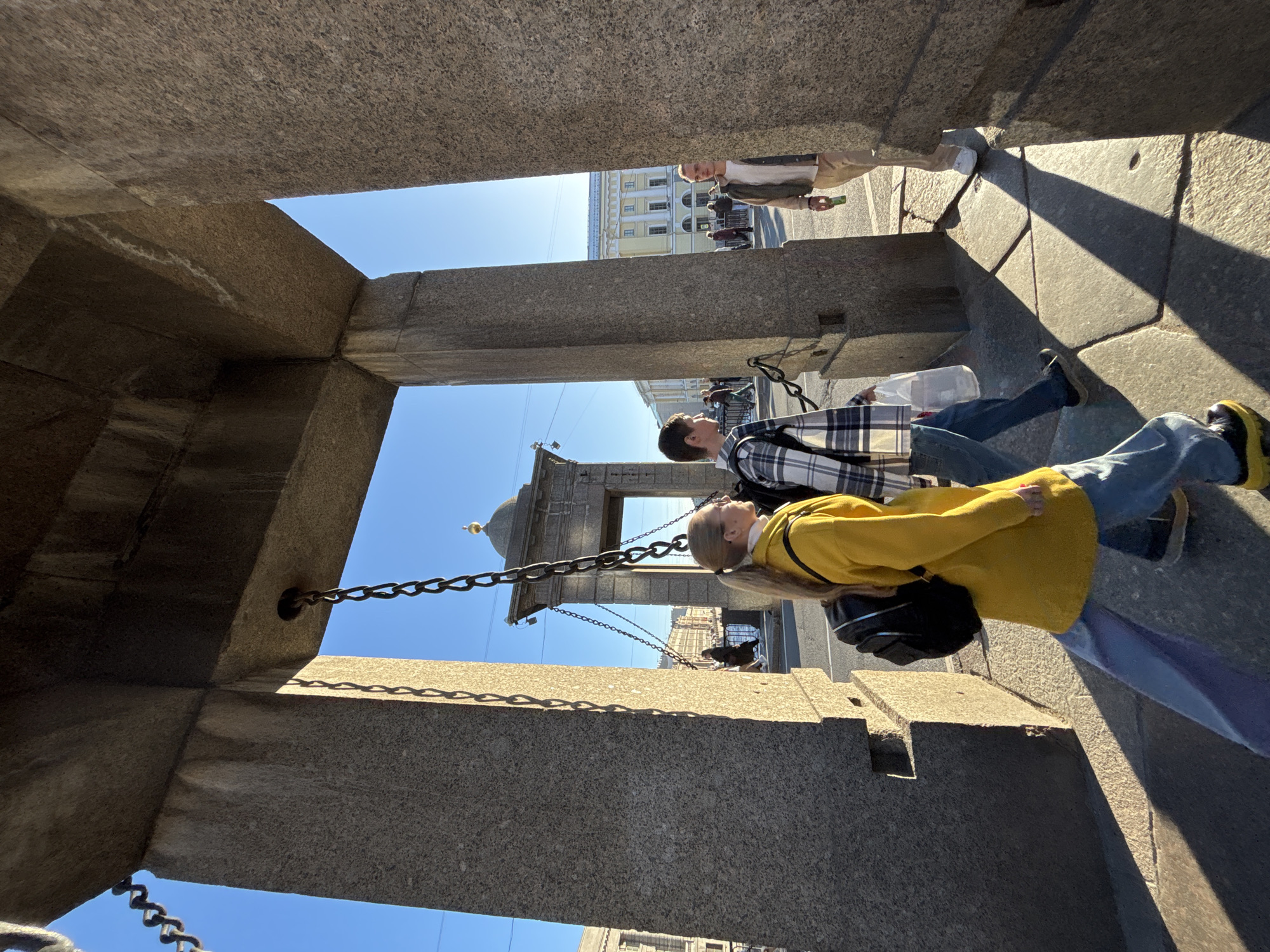 The image depicts a view through a stone structure, revealing a street scene in Saint Petersburg, Russia. The structure appears to be a part of the arch of the General Staff Building on Palace Square, as suggested by the chains and architectural features visible.
Two people are prominent in the middle ground: one wearing a yellow sweater, blue jeans and carrying a black backpack, and another wearing a plaid shirt and blue jeans, both walking away from the viewer. A third person is walking towards the right side of the frame. Several other people are visible further down the street.
The background includes a building with a light-colored facade, typical of the architecture in Saint Petersburg. The sky is clear and blue. The ground in the foreground is made of stone slabs, with shadows indicating the time of day.