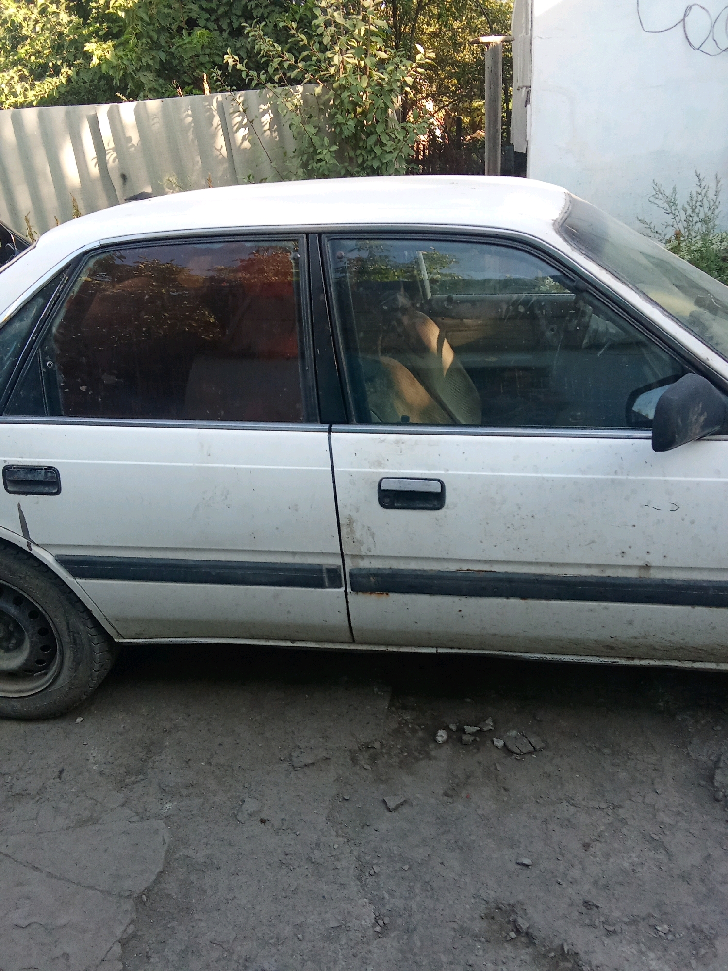 The image shows the side of a white, four-door sedan parked on a dirt or gravel surface. The car appears to be older and shows signs of wear, including dirt and some rust. The windows are tinted, and the interior is partially visible, showing a beige seat. A black side mirror is attached to the driver's side door, and a black door handle is also present. The car is parked next to a corrugated metal fence, behind which is dense green foliage. A white building with some scribbled markings is visible on the right side of the image. The ground is uneven and appears to be a dirt path or lot. The overall scene suggests an outdoor setting, possibly in a rural or semi-urban area. The location in Gavrilovka, Kyrgyzstan, is noted.