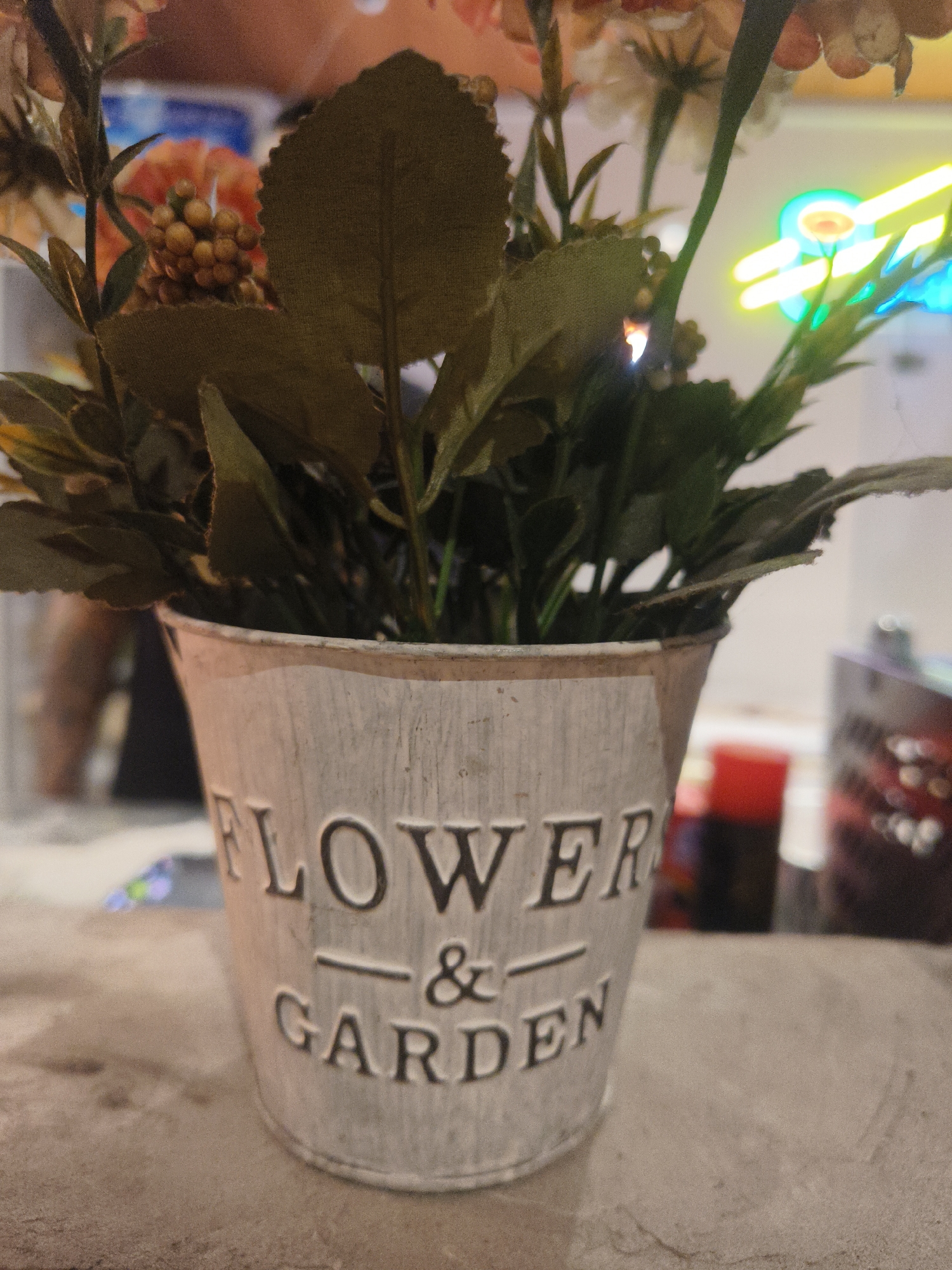 An artificial floral arrangement sits in a white, distressed metal pot with the embossed words "FLOWER & GARDEN" on its exterior. The pot is placed on a light brown, textured surface that appears to be a countertop or table. In the blurred background, a neon sign with bright yellow and blue lights is visible, suggesting an indoor setting possibly in a commercial establishment. The arrangement itself consists of green leaves, small clusters of berry-like structures, and some indistinct floral elements with hints of orange and pink. The overall scene is dimly lit, with the neon sign providing a contrasting splash of color. The image does not contain any identifiable people or specific landmarks that would allow for a definitive location within Kuta Selatan, Indonesia.