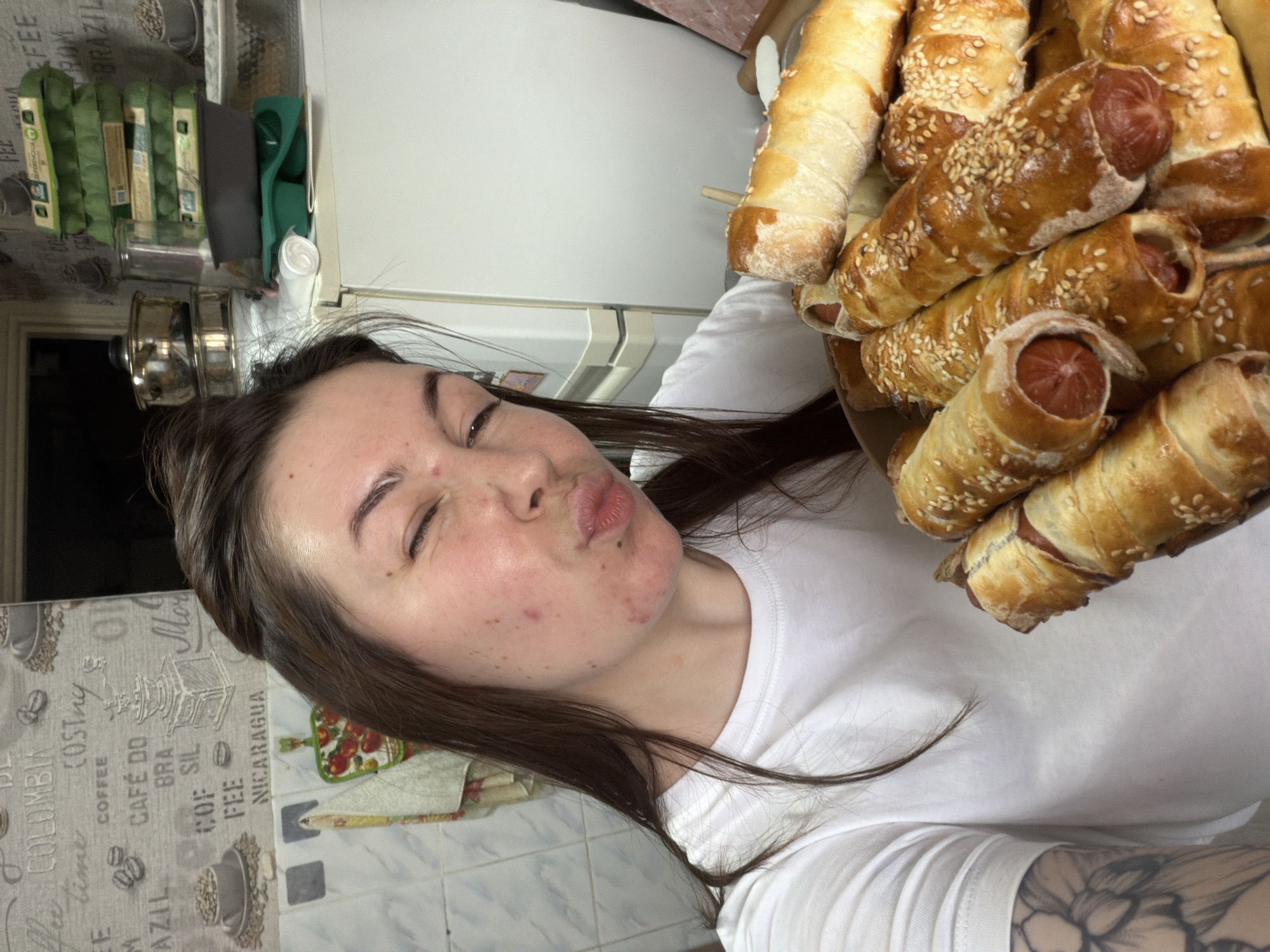 A woman with long brown hair, wearing a white T-shirt, holds a pile of sausage rolls close to her face. The pastries are golden brown and sprinkled with sesame seeds. The woman has a pouting expression and one eye closed. The background includes a kitchen counter with coffee-themed decor, a glimpse of a refrigerator, and various kitchen items like egg cartons and a stainless steel pot. The location is a kitchen, which could be in the city of Chaikovsky, Russia.