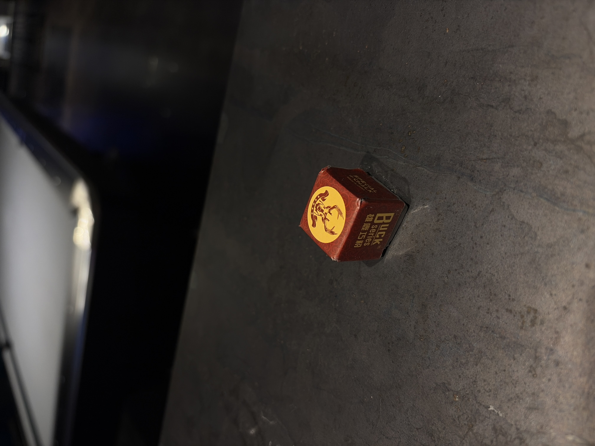A single, square piece of chalk rests on a dark, matte surface. The chalk's packaging is red and features a gold-colored emblem of a deer head. The text "BUCK SERIES" is printed on the side in gold lettering, alongside other markings that appear to be in Chinese characters. The surface under the chalk shows slight variations in texture. To the left, a blurred, light-colored object is visible, possibly part of furniture or a fixture. The location is unknown.