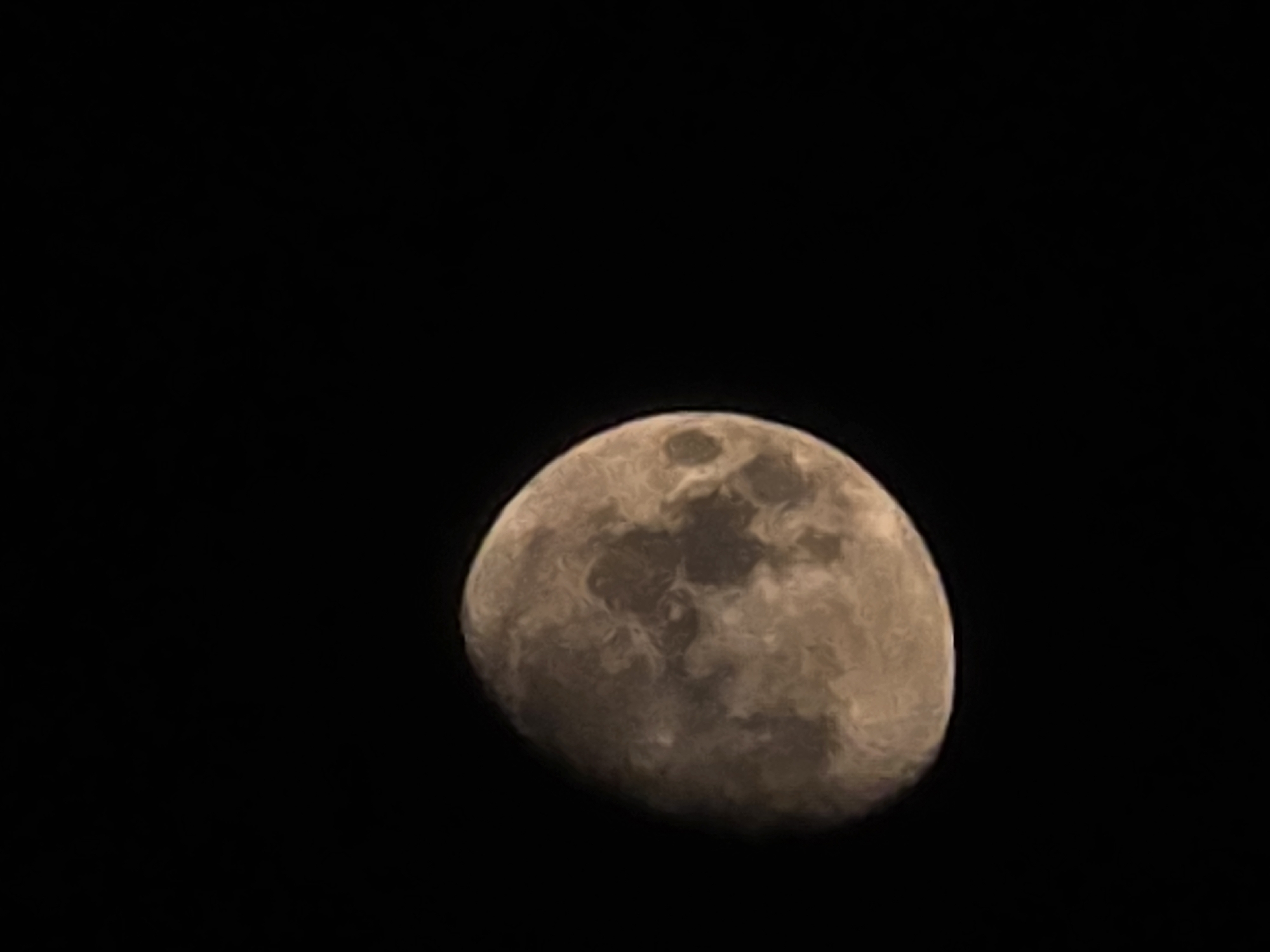 The image shows a close-up view of the moon against a black, starless sky. The moon is partially illuminated, with visible craters and surface features. It appears to be in a gibbous phase, with more than half of the moon lit. The colors of the moon range from light brown to dark gray, indicating variations in its surface composition. The image does not contain any elements that would allow for the identification of a specific location in Yambol, Bulgaria, or any other city. There are no people, objects, or actions depicted in the scene that provide contextual information about its setting.