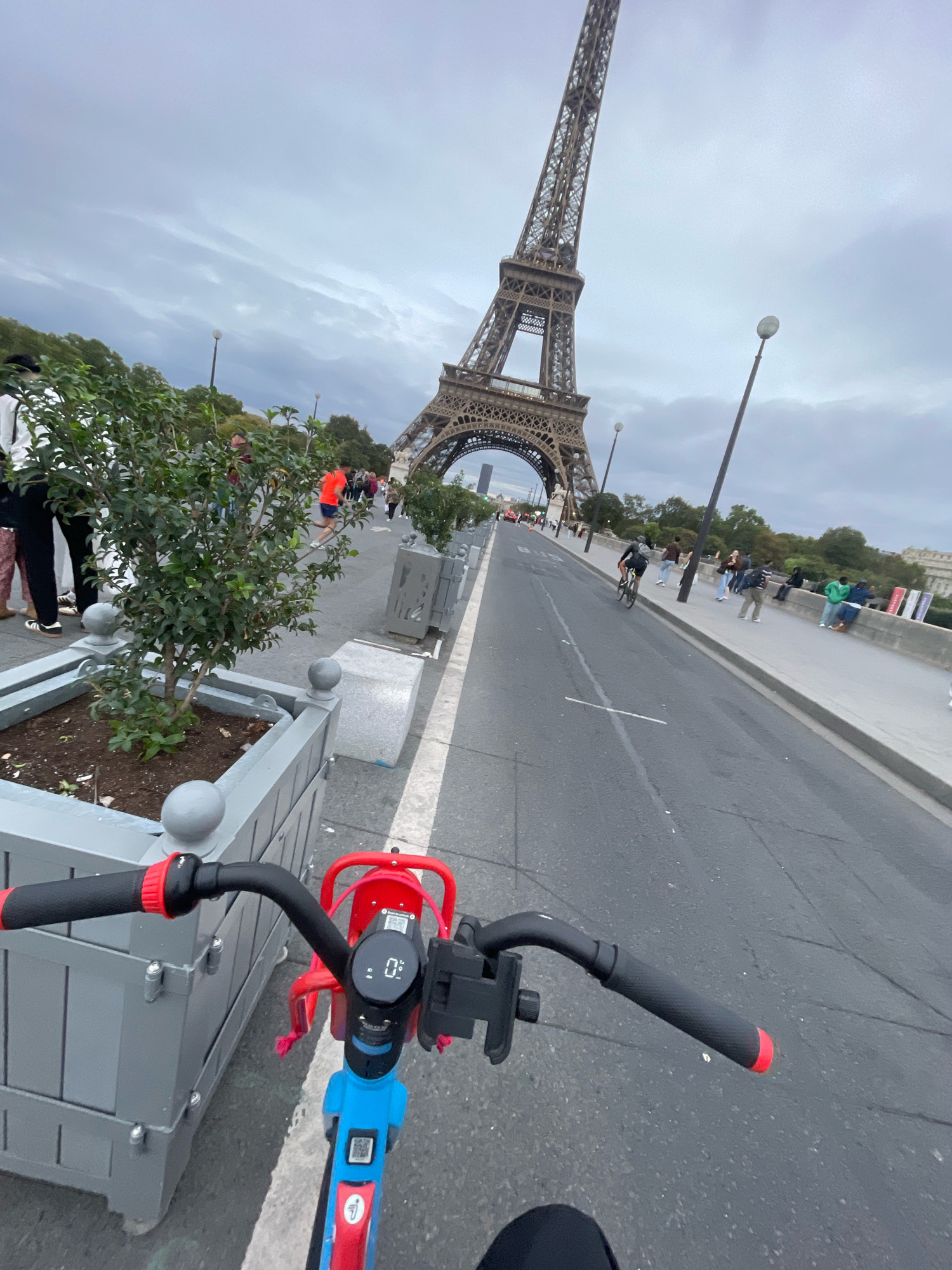 An over-the-shoulder perspective, likely from a person riding a blue and red electric scooter, shows the Eiffel Tower dominating the background under a cloudy sky. The scooter's handlebars, display screen showing "0 km/h", and part of its frame are in the foreground. To the left, gray planters with small trees are lined up along a walkway. People are visible on the walkway, some walking, others sitting, and a cyclist is in the distance on the road. The scene is set in Paris, France, likely on a bridge or promenade near the Eiffel Tower.