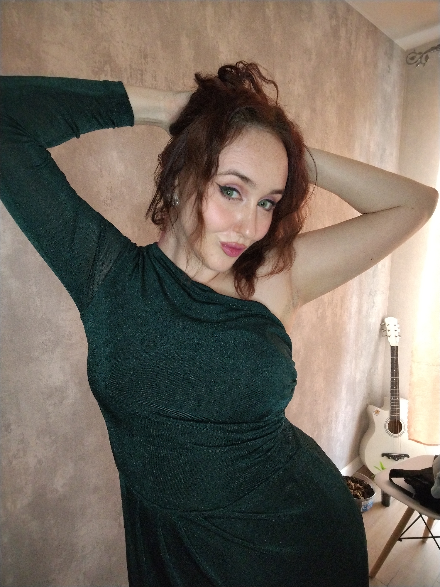 An indoor portrait-style photograph, captured in Izhevsk, Russia, features a woman posing confidently. The subject is a woman with reddish-brown, curly hair styled loosely on top of her head, striking green eyes, pink lipstick, and visible freckles across her forehead. She wears a dark green, shimmery, one-shoulder dress; the left arm features a long sleeve with a sheer mesh detail near the shoulder. Her left arm is raised and bent behind her head, while her right arm is also raised with her hand near the back of her head, revealing some natural underarm hair. She looks directly at the viewer with a subtle, engaging smile.
The setting appears to be a room in a home. The walls are covered in a light beige or light brown textured wallpaper. To the right side of the frame, a white acoustic guitar with a black pickguard rests against the wall. Adjacent to the guitar is a small, minimalist chair with a white seat and light wooden legs, upon which a dark object, possibly a bag, is placed. On the floor between the chair and the guitar, a plastic container holds soil and some dry plant matter. Soft, natural light illuminates the scene, suggesting it is daytime. No text is visible in the image. The woman's pose and expression convey confidence and a playful demeanor within her personal space.