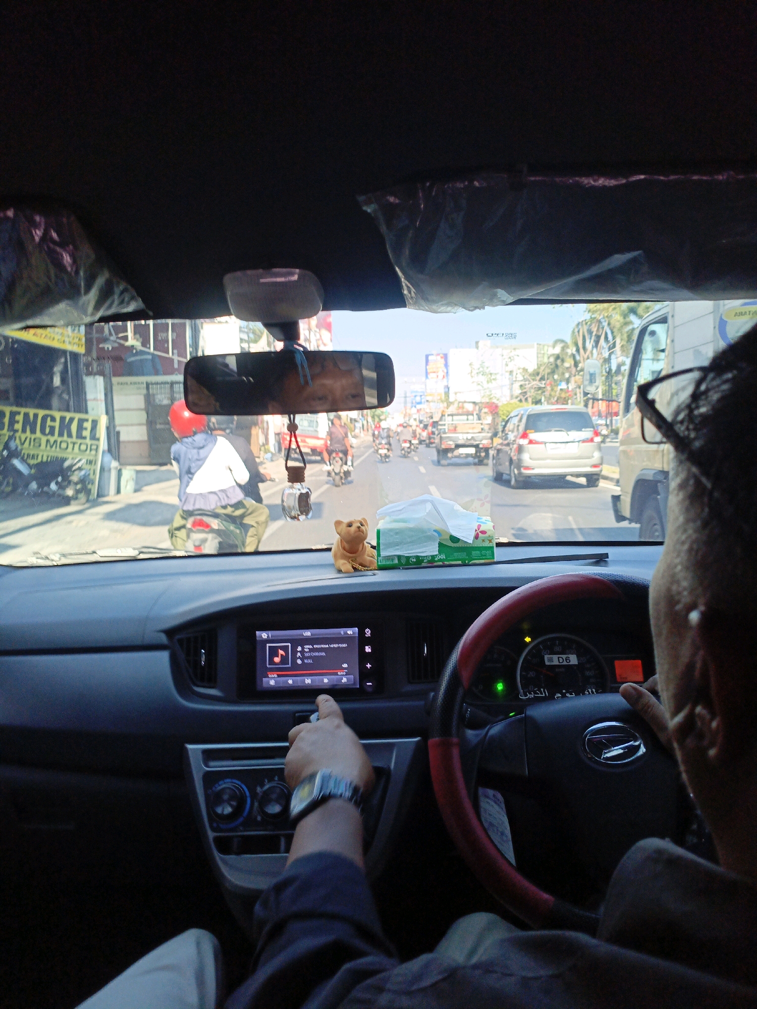 The image is taken from the interior of a car, looking forward through the windshield. The view captures a street scene in Sidoarjo, Indonesia, on a sunny day. The car's dashboard is visible in the foreground, with a touchscreen infotainment system displaying music controls. A hand wearing a watch is reaching out to touch the screen. To the right of the dashboard is a steering wheel with the brand logo. The windshield shows traffic, including motorcycles, cars, and a truck. A billboard with the text "BENGKEL MOBIL" (Motorcycle Workshop) is visible on the left side of the street. A small, tan teddy bear figurine sits on the dashboard. The overall impression is of a busy urban environment during daylight hours.