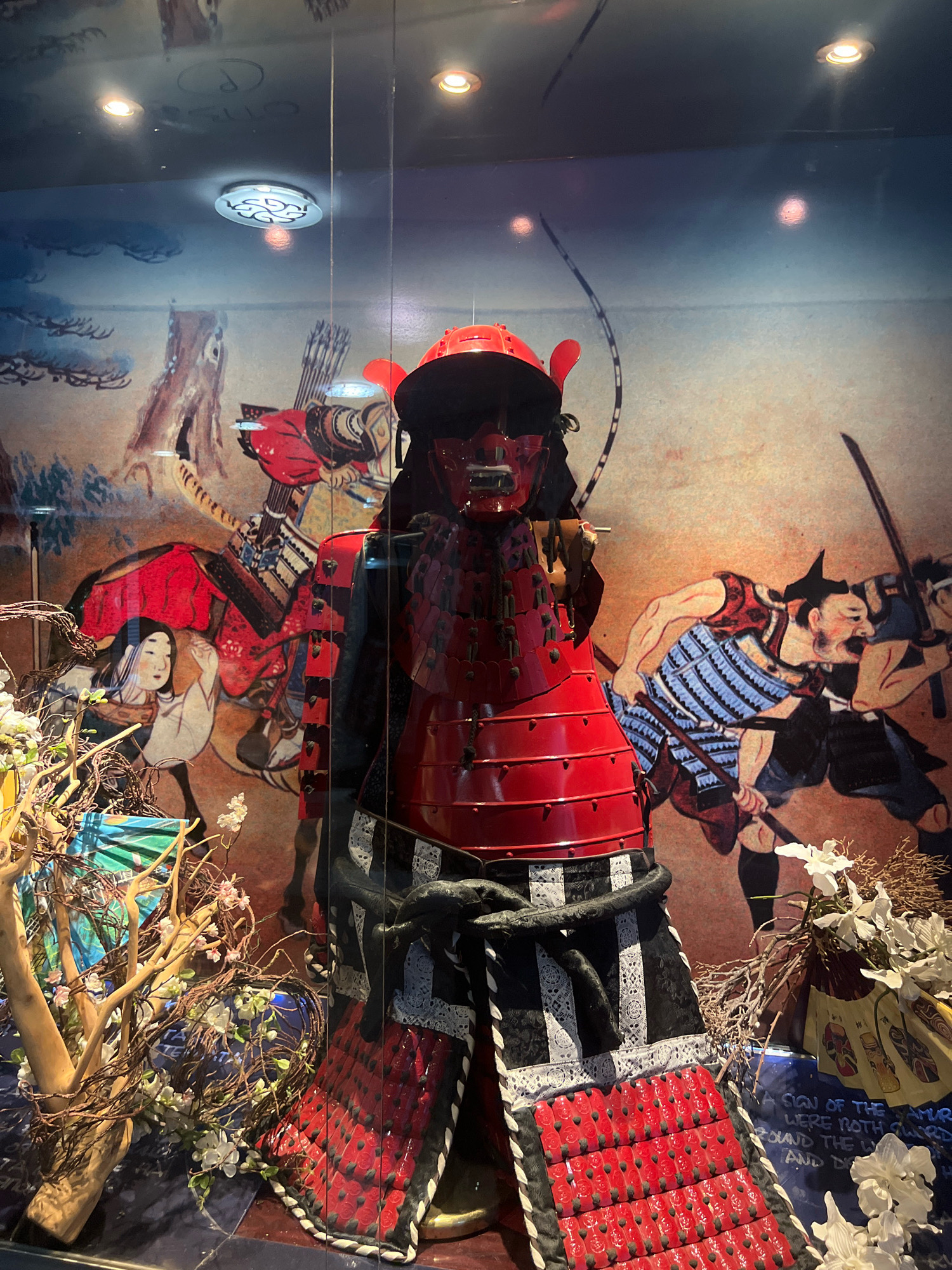 The image shows a display of traditional Japanese samurai armor within a glass enclosure, likely in a museum. The armor is predominantly red with black and white accents. Behind the armor is a mural depicting scenes of samurai combat and figures, rendered in a traditional Japanese art style. In the foreground, there are decorative branches with white and pink flowers, and some blue fabric. The lighting in the enclosure includes spotlights on the ceiling. A sign with text is partially visible at the bottom right. This display is located in Sofia, Bulgaria, specifically at the National Museum of Military History.
