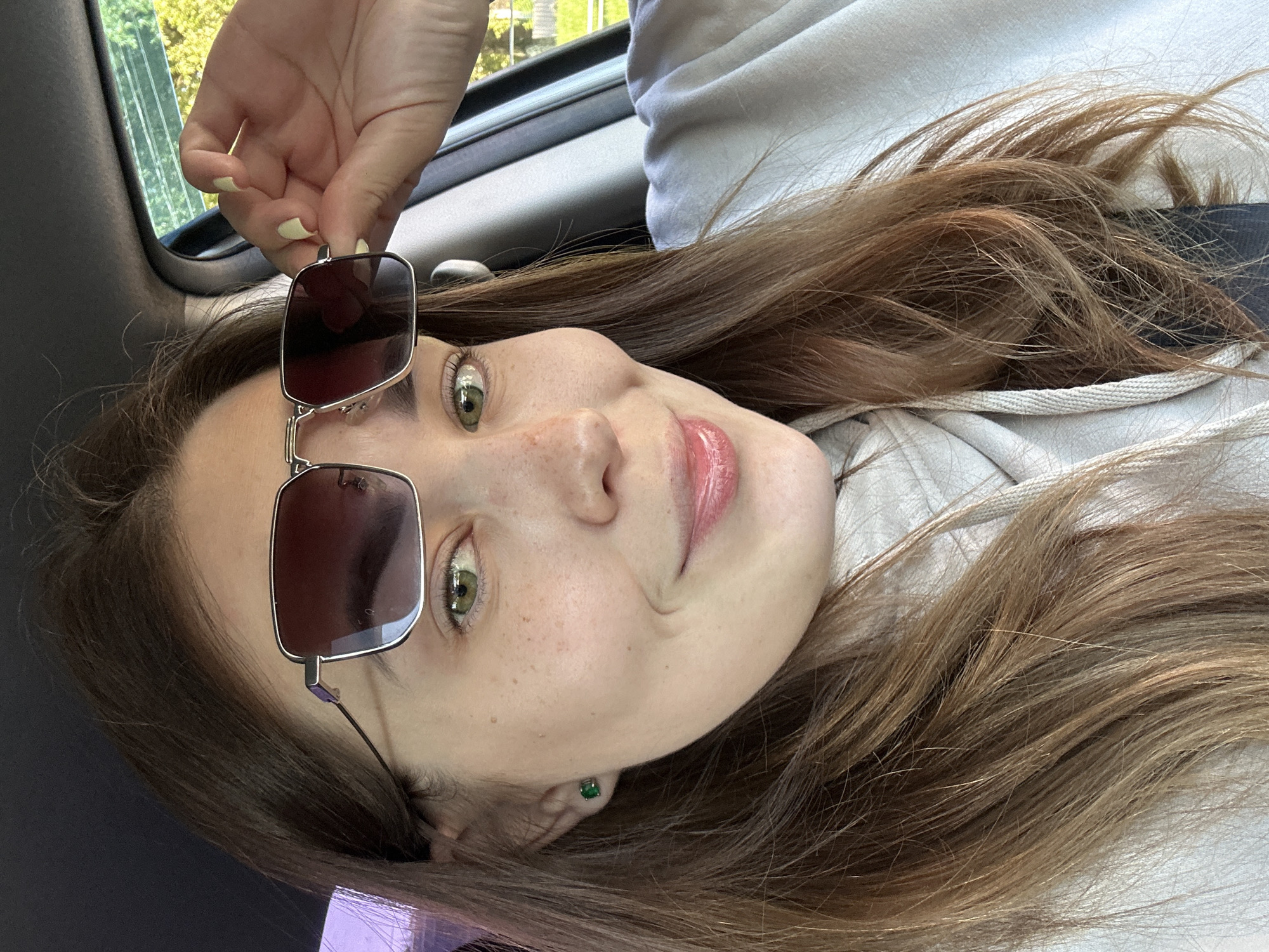 A young woman with long brown hair and green eyes is sitting in a car and holding her sunglasses up to her head with her right hand. She is wearing a light-colored hoodie and a green stud earring in her left ear. The car appears to be parked outdoors, with a blurred background of green foliage and trees, suggesting a location in a suburban or rural area. The lighting indicates it is daytime. There are no visible landmarks that would specifically place this image in Perm, Russia.