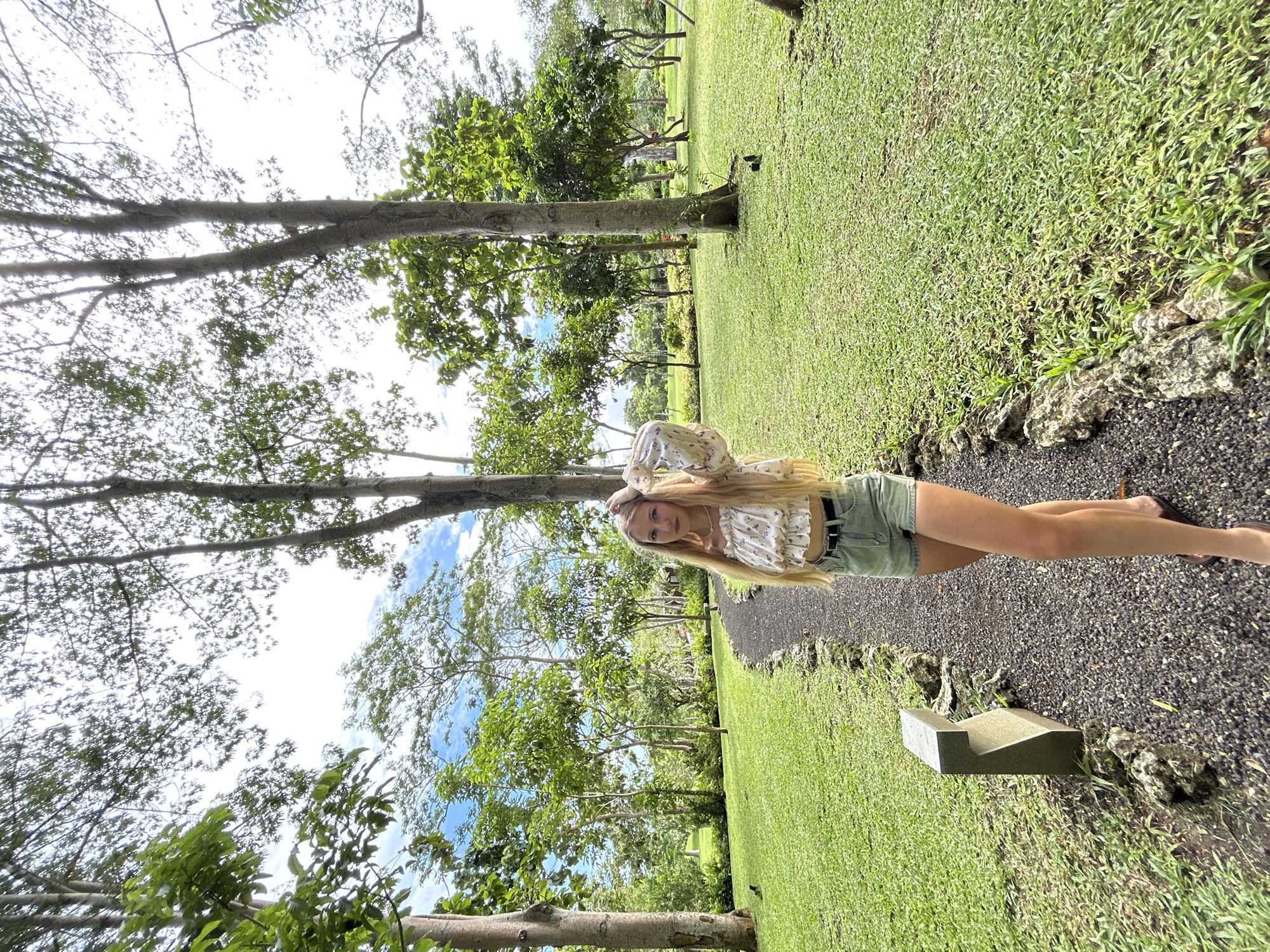The image displays a serene outdoor scene within a lush park or garden, consistent with the tropical environment of Kuta Selatan, Indonesia. The main subject is a young woman with long blonde hair, standing on a dark gravel path. She is wearing a light-colored, ruffled top with a subtle floral pattern, light denim shorts with a dark belt, and flip-flops. Her gaze is directed towards the camera with a neutral, calm expression, and one hand is resting near her head, suggesting she is posing for a photograph.
The gravel path is bordered by natural rocks that separate it from expansive, well-maintained green lawns on either side. Numerous trees with light-colored trunks and dense green foliage line the path and fill the background, creating a verdant setting. The sky above is partly cloudy, indicating bright daytime conditions without harsh sunlight. To the right of the path, a low, dark grey, rectangular object, possibly a stone bench or marker, is visible on the grass. There is no visible text in the image. The scene conveys a peaceful and natural ambiance.