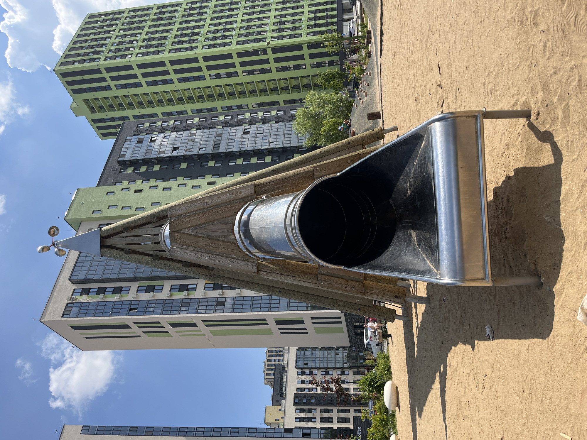 In a sandy playground, a large, conical wooden structure with a silver metal slide extending from its peak dominates the foreground. The slide's opening is wide and curved, with a polished metal finish. Behind the playground, several modern, tall buildings with colorful facades—some green and black, others beige and green—rise against a bright blue sky with scattered white clouds. In the middle ground, trees and shrubs create a line of greenery separating the playground from the buildings. Several people are visible, including two individuals seated on a bench near the slide and a child playing on a mound in the background. The scene appears to be a contemporary urban residential area with recreational facilities.