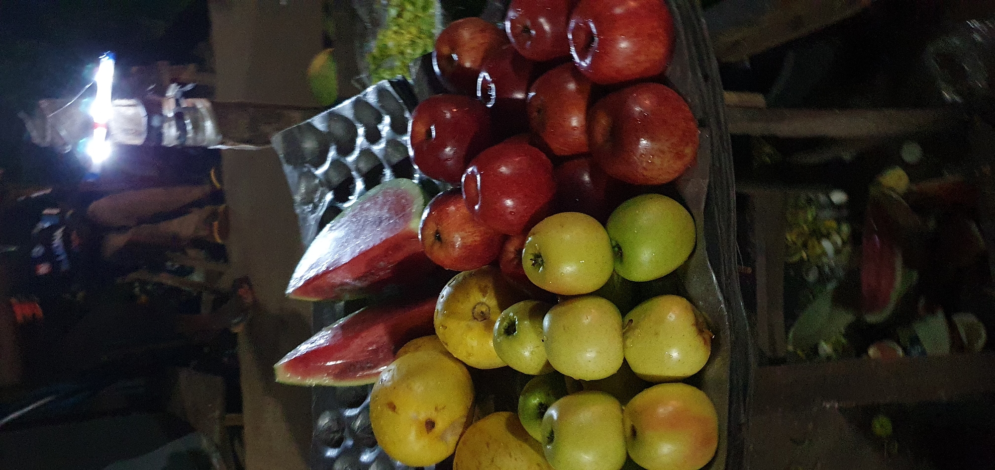 The image depicts a stall in Barakin Angundi, Nigeria, at night. The stall is laden with various fruits, including several slices of watermelon, yellow fruits that appear to be guavas or similar, and numerous red and green apples. The fruits are arranged on a dark surface, possibly a table or tray, with a textured, possibly metallic, object behind the watermelon slices. In the background, several individuals are visible, partially obscured by darkness. One person is standing and appears to be illuminated by a bright, possibly handheld, light source. The overall scene suggests a bustling nighttime market environment, with the fruits illuminated by artificial light.