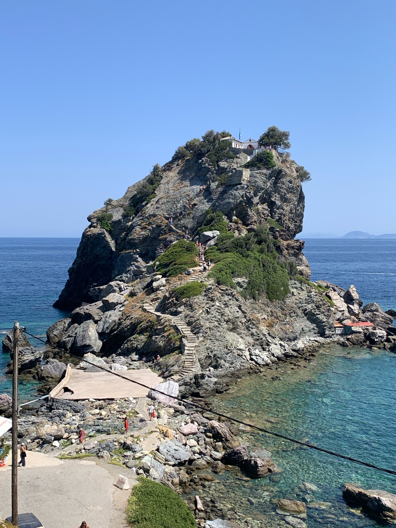 This is a photograph of the Agios Ioannis Church, also known as the Mamma Mia Chapel, located on a rocky outcrop near the town of Glossa, Skopelos, Greece. The scene is dominated by the imposing rock formation rising from the clear blue sea. A white chapel with a red roof and a Greek flag is situated at the summit of the rock. A winding stone staircase, with numerous steps, ascends the side of the rock, and several people are visible on it, either climbing up or down. Lush green vegetation clings to the rocky slopes.
In the foreground, there is a rocky shoreline with a concrete platform. Several people are present on this lower level, some walking along paths, others gathered on the platform. The water is crystal clear, revealing the rocky seabed below. A utility pole with wires is visible on the left side of the image. The sky is a clear, cloudless blue. The overall environment is a sunny, coastal landscape.