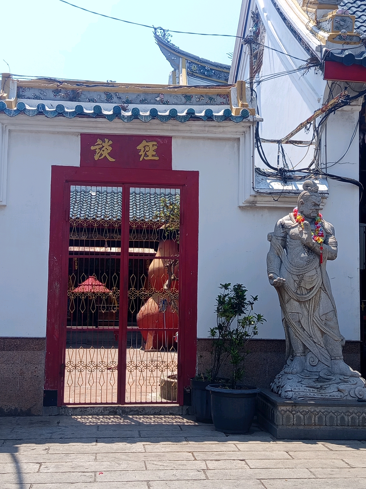This image shows the entrance to a temple in Semarang, Indonesia. The temple has a red door with ornate gold lettering above it. To the right of the door, there is a stone statue of a warrior wearing a flower garland. The statue stands on a decorative pedestal. In front of the temple, there is a paved walkway. The sky is clear and blue, suggesting it is a sunny day.