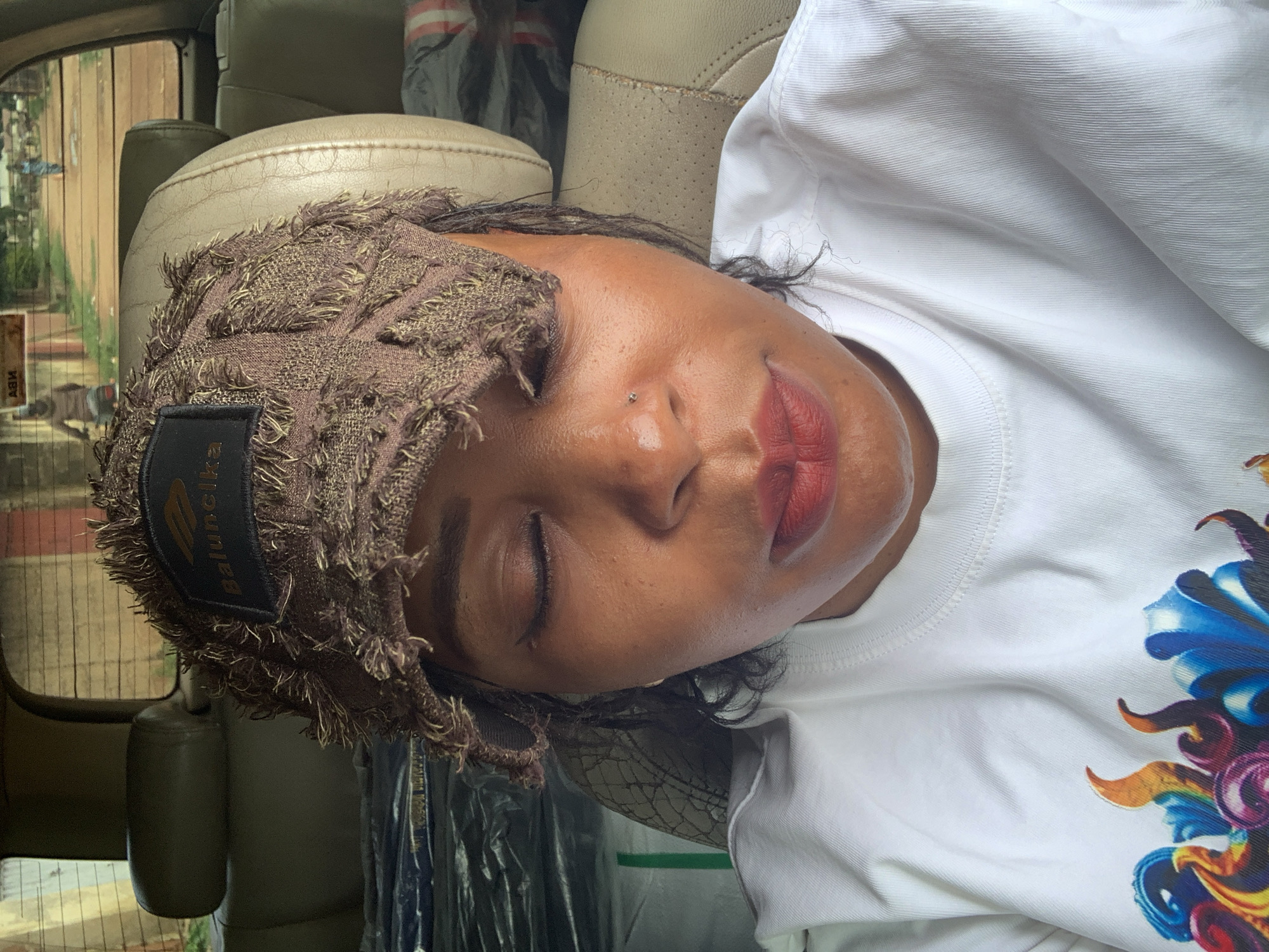 A medium close-up shot shows a woman from the chest up, sitting in the front seat of a car. She is wearing a distressed brown cap with a black patch bearing the text "Balunelka" and a white t-shirt with a colorful graphic print on the lower left side of the frame. Her eyes are closed, and her lips are parted slightly, with red lipstick applied. A small nose stud is visible on her right nostril. The interior of the car, with beige leather seats and some luggage in the background, is visible. Through the rear window, a dusty street in Sabon Tasha, Nigeria, is depicted, with people and a motorcycle in the distance. The overall lighting suggests it is daytime.