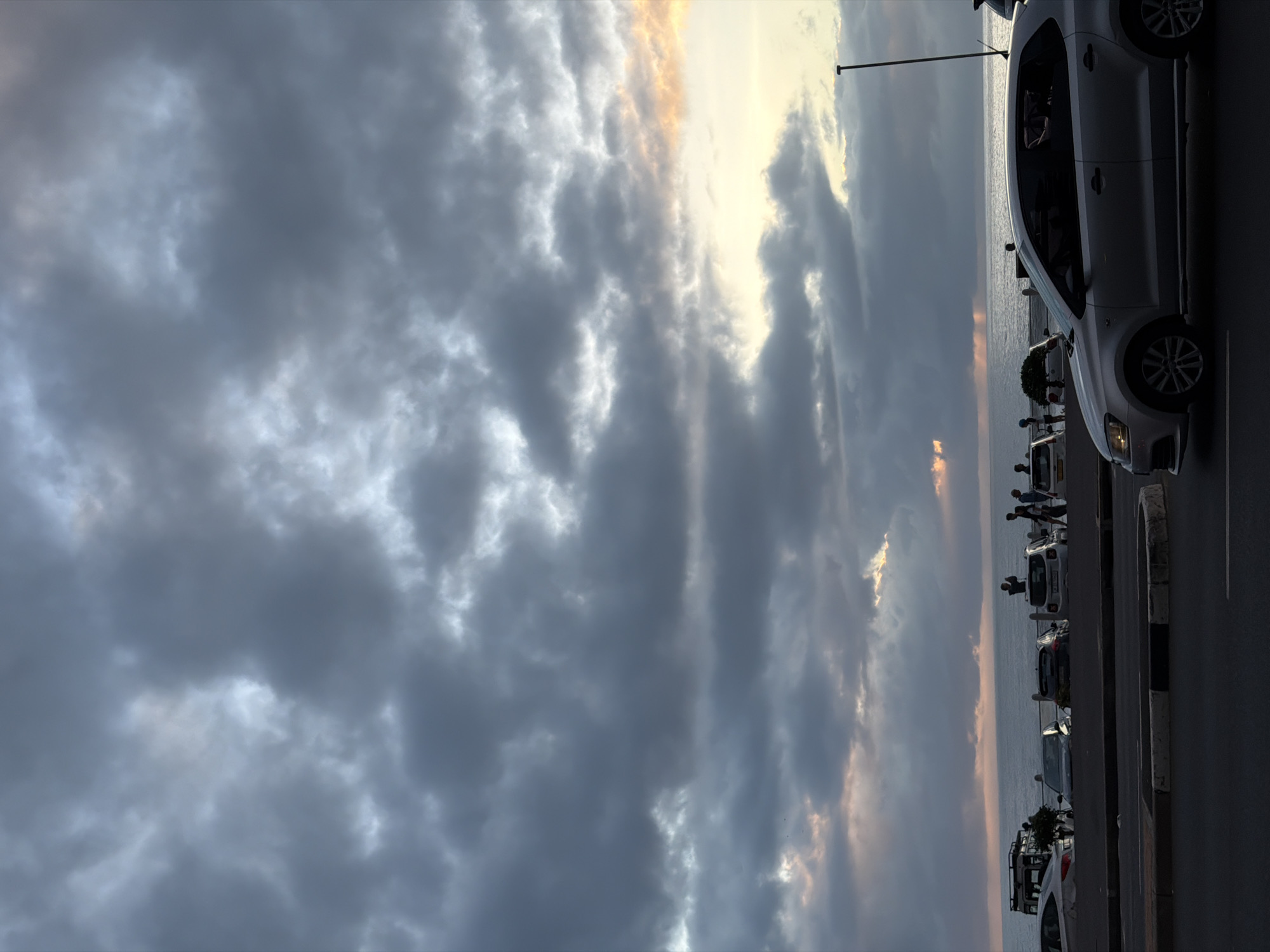 The image shows a street in Cape Town, South Africa, alongside the ocean. Cars are parked along the side of the street, with people standing and looking out at the ocean. The sky is cloudy, with the sun peeking through the clouds.