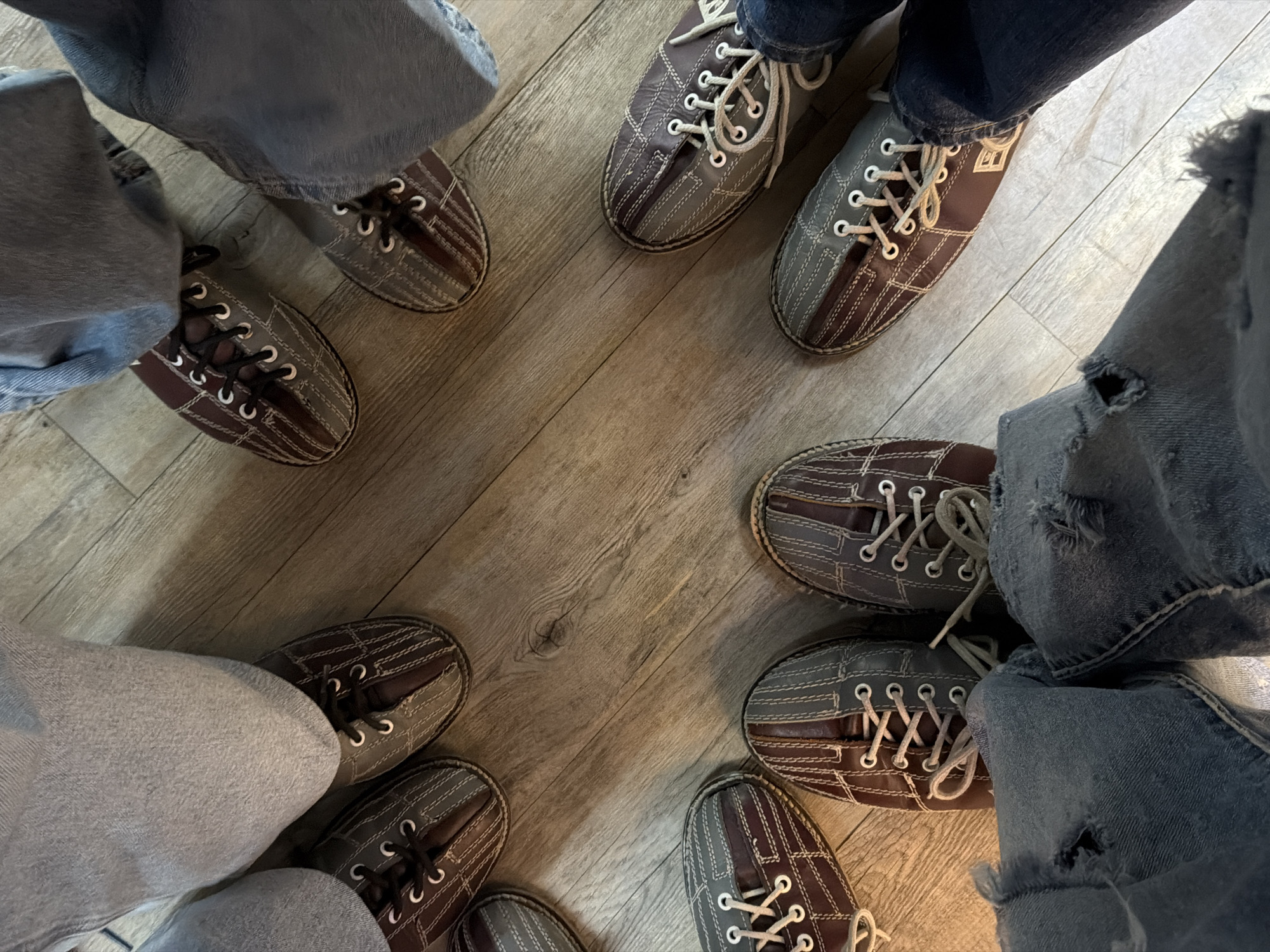 An overhead view captures the lower legs and feet of at least six individuals standing on a light-colored wooden floor, characteristic of a bowling alley. Each person wears a pair of two-toned bowling shoes, combining brown/burgundy and grey/blue-grey sections with prominent white or light-colored laces and eyelets. The shoes feature decorative stitching patterns. Visible above the shoes are the lower portions of diverse denim jeans, including light and dark washes, with some displaying ripped details at the cuffs. The feet are positioned in a loose circular arrangement, oriented towards the center of the image. Shadows are cast by the individuals onto the wooden surface, indicating overhead lighting. The scene takes place within a bowling establishment, such as Bol Magia Puebla, located in Puebla, Mexico.
