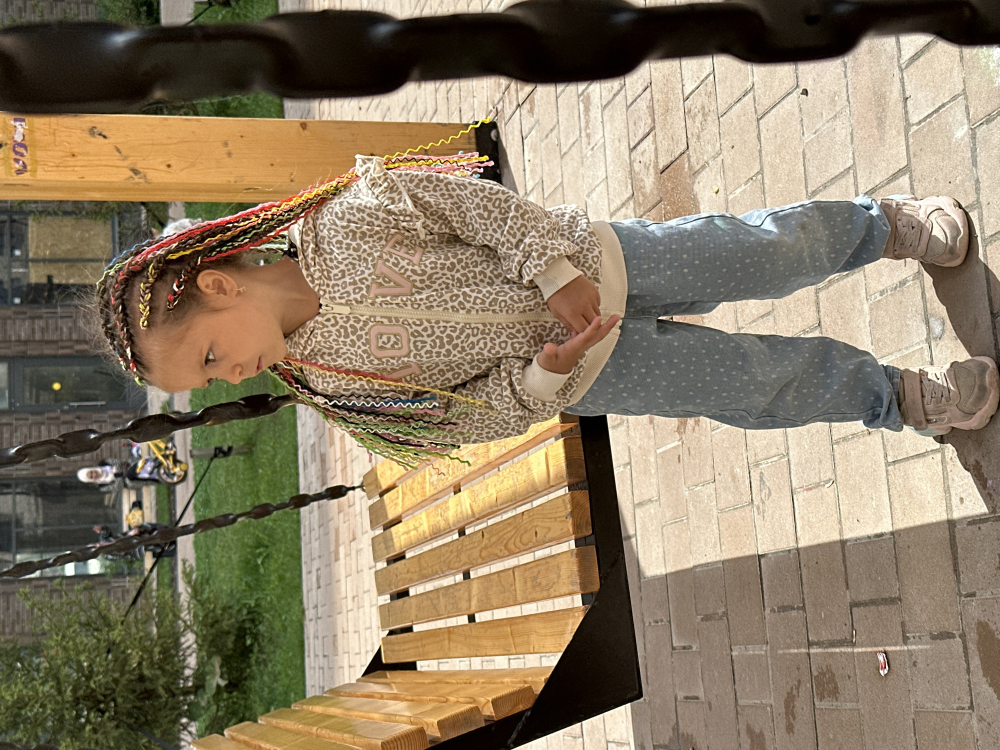 A young girl with brightly colored braided hair stands in front of a wooden swing set. She is wearing a leopard-print hooded jacket and light blue dotted pants. The girl is looking to her left, with her hands clasped in front of her. The swing set is made of wood and has black metal chains. The ground is paved with brick-like stones. In the background, a grassy area is visible, with some trees and buildings. There is also a glimpse of a yellow and black motorcycle with a person on it, and another person in the background wearing a white headscarf. The scene appears to be an outdoor playground or park.