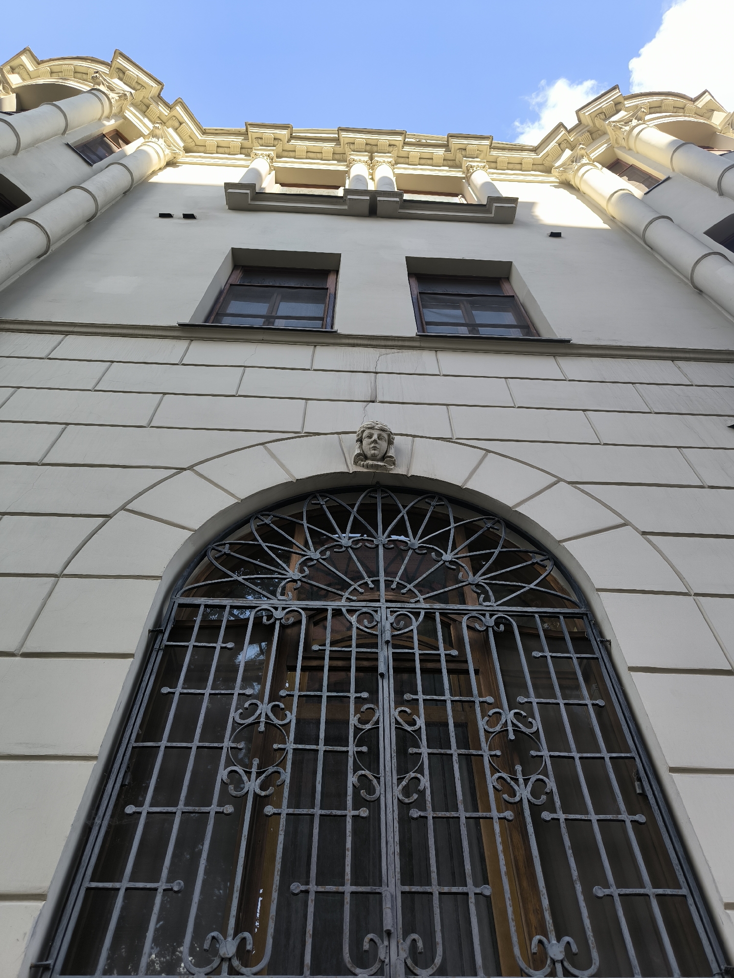 An exterior, low-angle, wide shot captures the ornate architecture of a building against a clear blue sky with scattered clouds. The building's facade is a light cream color, with a textured stone base divided into rectangular blocks. Above this, a large, arched window is covered by a decorative wrought-iron grate featuring intricate scrollwork and a circular motif in the center. The window itself appears to have wooden panes and is tinted dark. A small, carved stone face is embedded in the keystone of the arch. Higher up on the building, two rectangular windows with wooden frames are visible. The top of the building features classical architectural elements, including columns, cornices, and decorative moldings, all rendered in a lighter, yellowish hue. The building is bathed in sunlight, casting subtle shadows. The scene is devoid of people or any signs of activity.