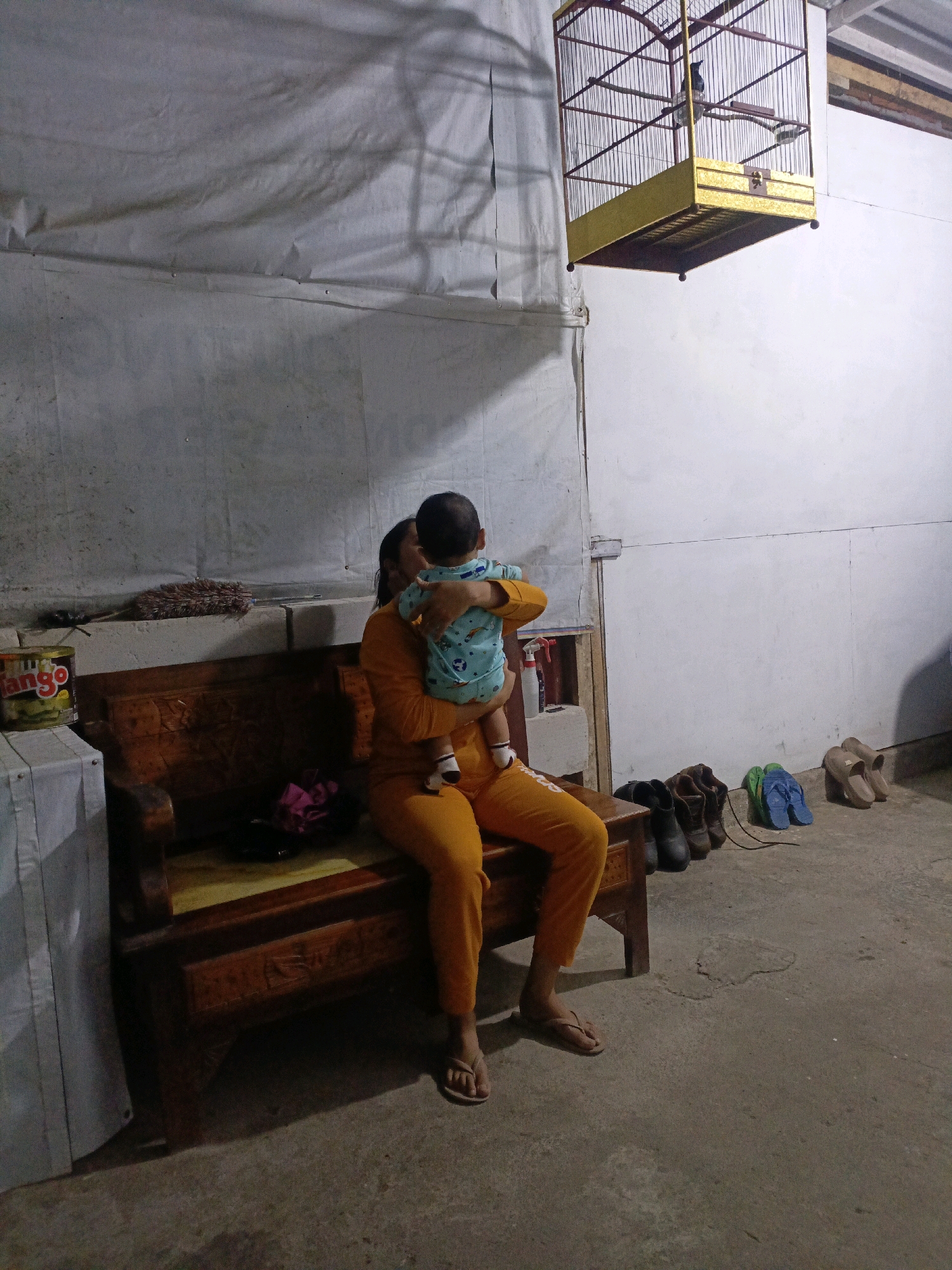 An indoor scene is depicted, likely a rural residence in Pager, Indonesia. In the foreground, a woman wearing an orange outfit is seated on a wooden bench, holding a baby. The baby is dressed in a light blue outfit with green accents. To the left of the bench, there is a cylindrical container with "jengo" written on it. Above and to the right of the bench, a yellow birdcage is hanging from the ceiling, containing a small black bird. The background consists of white walls with a fabric draped across the top, creating shadows. On the floor to the right, several pairs of shoes and flip-flops are arranged. The environment appears somewhat rustic and domestic.
