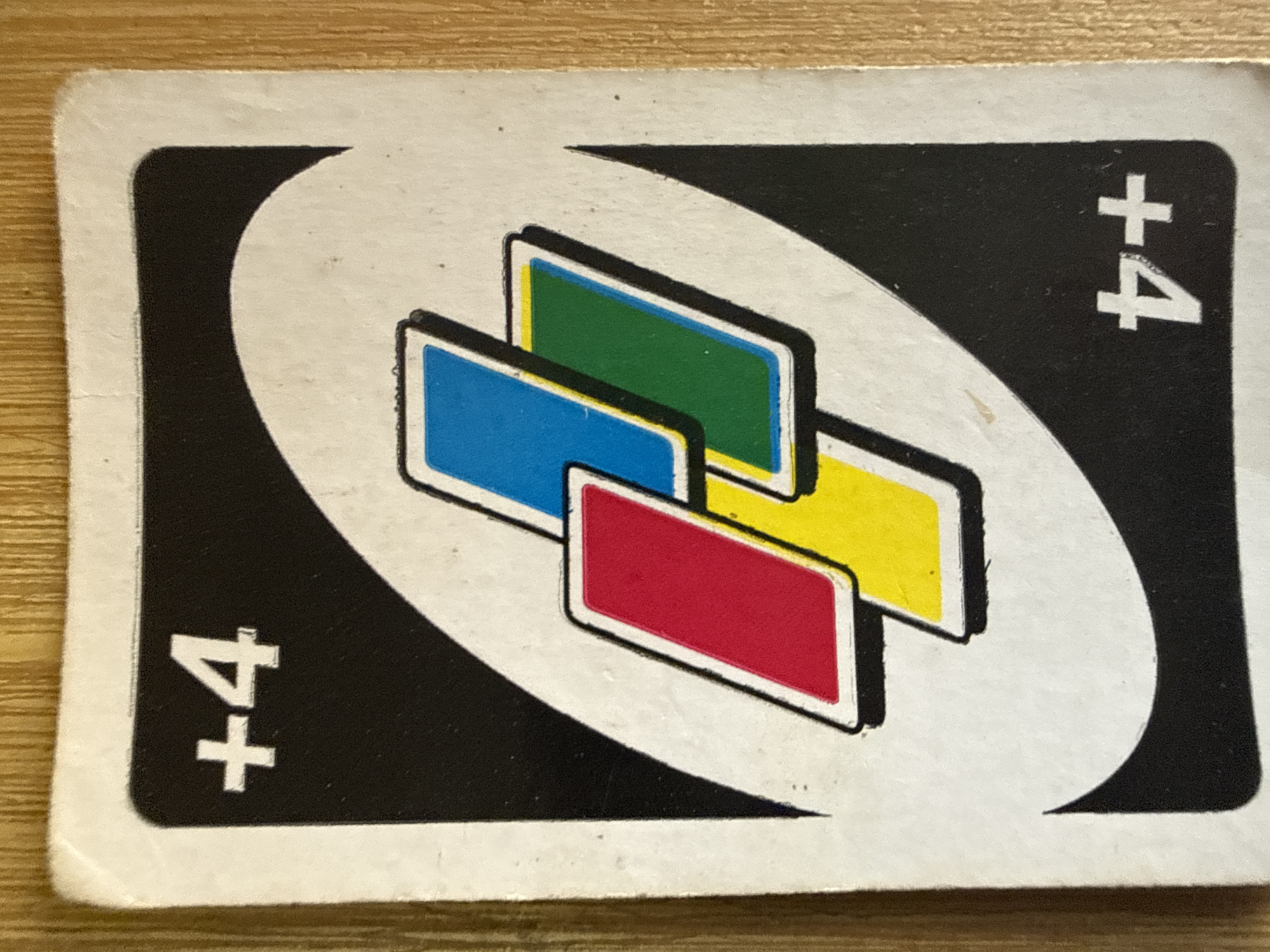 The image displays a close-up of a single Uno card, specifically a "+4" card. The card has a white border surrounding a black background. In the upper left and lower right corners, the "+4" symbol is prominently displayed in white. The central design features a stylized representation of four colored Uno cards overlaid on each other. The colors visible are red, blue, green, and yellow, each depicted with a white border and a black outline. The card is placed on a wooden surface, likely a table, with visible wood grain. There are no people, actions, or interactions depicted in the image. The location in Tbilisi, Georgia, cannot be determined from the provided image.