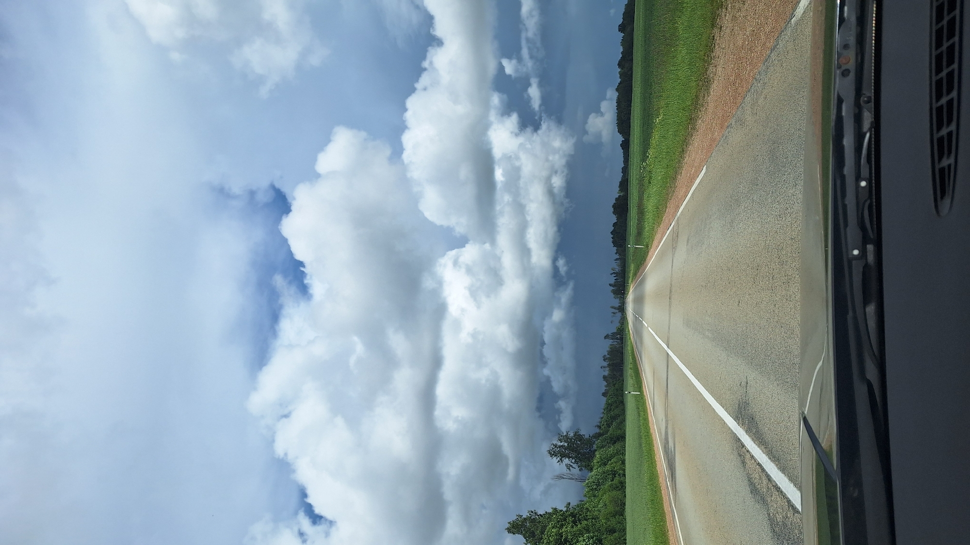 The image depicts a view from the driver's seat of a car on a two-lane asphalt road in an unidentified location in Latvia. The road stretches into the distance, flanked by green fields and trees. The sky is partly cloudy with dramatic, dark storm clouds gathering on the horizon, contrasting with patches of blue sky and bright white clouds. The dashboard and windshield wipers of the vehicle are visible in the foreground. There are no people or animals clearly visible in the image.