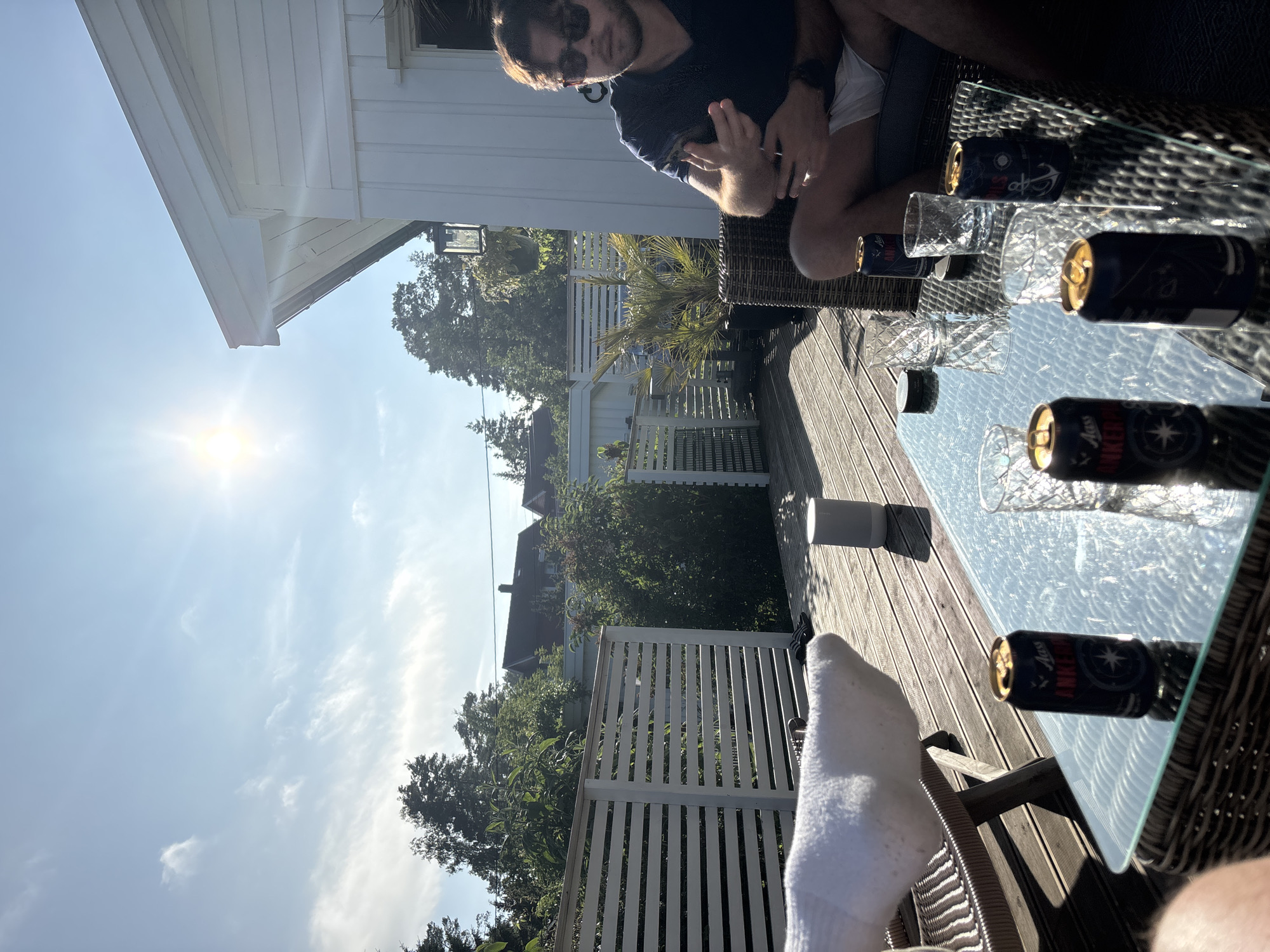 In Larkollen, Norway, on a sun-drenched wooden deck, three men are enjoying a relaxed outdoor gathering. The scene is set against the backdrop of a white house with horizontal siding, typical of Scandinavian architecture. A bright sun shines from the upper left corner of the frame, casting long shadows across the deck and glinting off the glass table.
In the foreground, a glass-topped coffee table is laden with four dark blue beer cans, each bearing the "Aass AnkerPils" label and a compass rose graphic. Two clear, patterned glasses sit near the cans, one appearing partially filled. A small, dark circular object, possibly a coaster or ashtray, is also on the table.
To the left of the table, a man's legs are visible, covered in white socks and resting on a wicker chair. A white terry cloth towel is draped over the armrest of this chair. Further back, a cylindrical white speaker sits on the deck.
To the right, a man with dark hair and sunglasses is seated on a wicker chair, wearing a dark blue t-shirt and white shorts. He is looking towards the table, with his right hand gesturing, index finger extended as if making a point or counting. Another man is partially visible behind him, also seated.
The deck is bordered by a white horizontal slat fence, behind which lush green trees and bushes are visible. In the distance, houses with red roofs are nestled amongst the trees, suggesting a residential area. The sky is a clear blue with scattered white clouds, and the overall atmosphere suggests a warm, pleasant day.